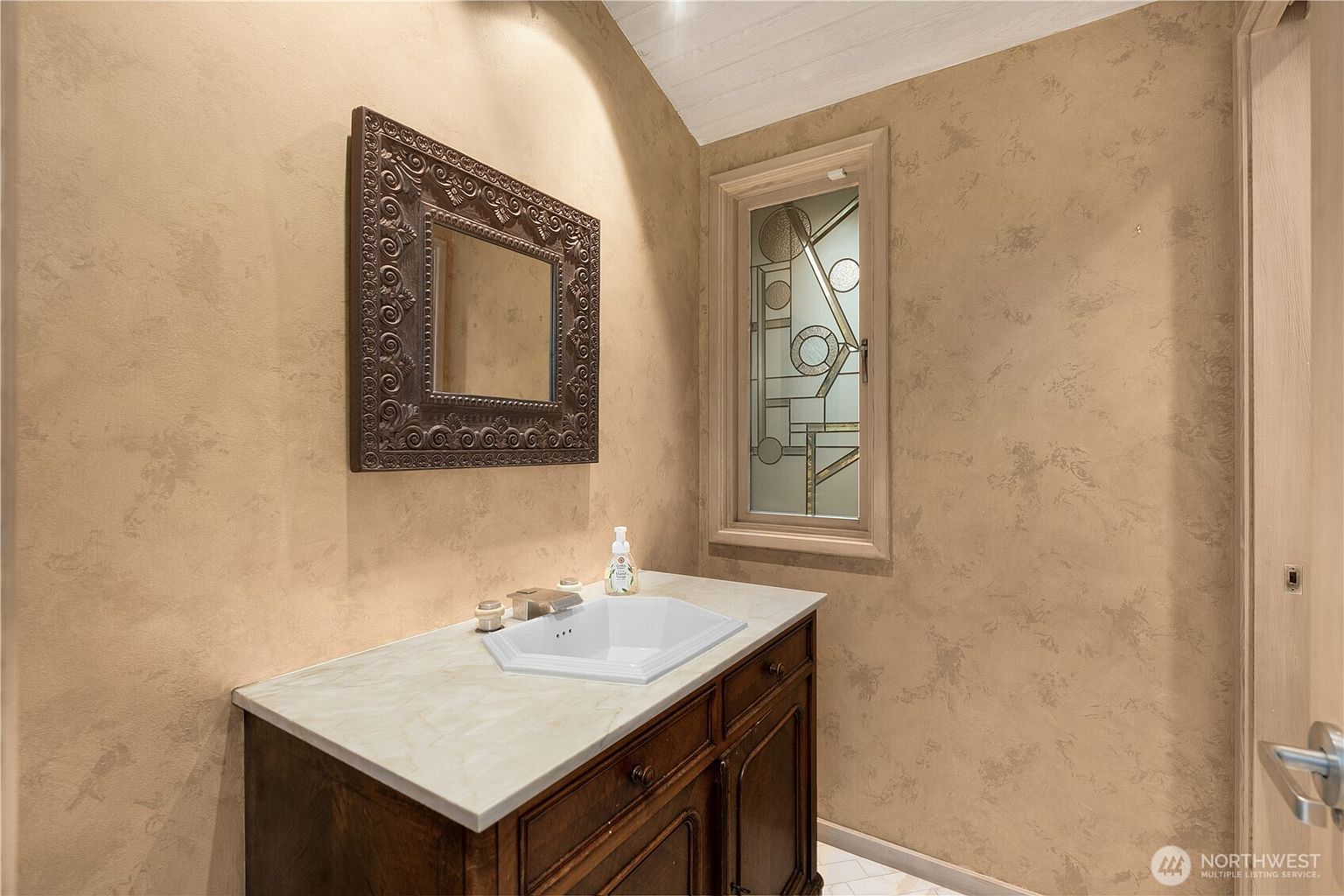 This powder room features a dark wood vanity topped with a light-colored stone counter and a unique geometric-shaped sink. The walls are finished in a textured, warm-toned faux paint, complemented by an ornate, dark-framed mirror and a tall, narrow window with decorative leaded glass. The space offers a sophisticated and intimate atmosphere, highlighted by the contrast between the dark cabinetry and the soft, neutral wall treatment.