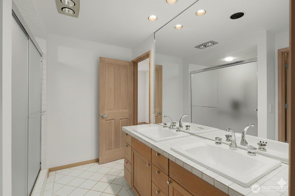 This bathroom features a dual-sink vanity with light wood cabinetry and a large, wall-spanning mirror that enhances the sense of space. The room includes a walk-in shower with a glass enclosure and light-colored tile flooring laid in a diagonal pattern. The perspective is from the doorway, capturing the functional layout and clean, neutral aesthetic of the space.