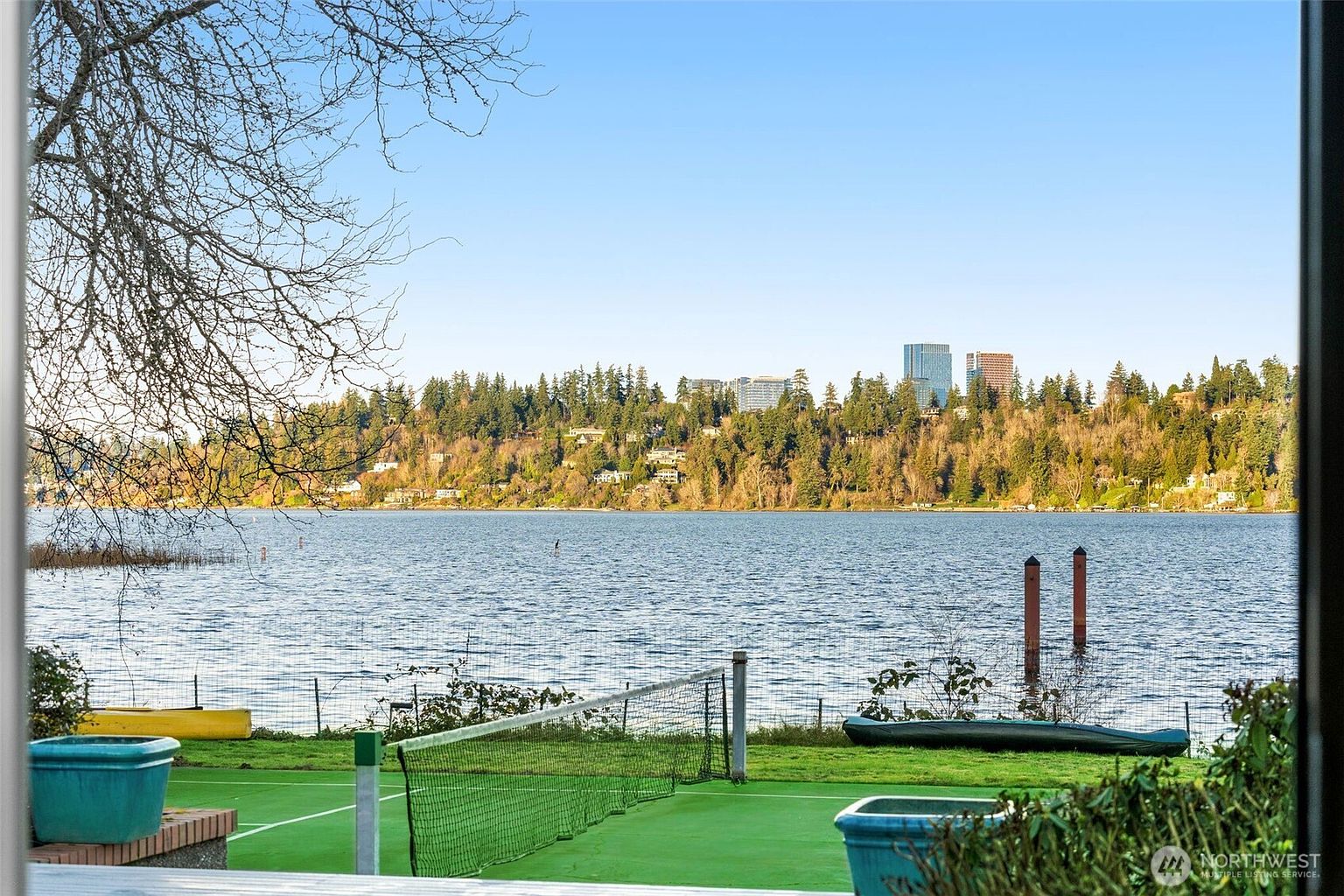 This view from a deck or patio overlooks a private waterfront yard featuring a green tennis or sports court and a grassy lawn. The scene captures a serene lake with two wooden pilings in the water, framed by a lush, tree-covered hillside and a distant cityscape under a clear blue sky. A yellow kayak and a green canoe are visible near the water's edge, emphasizing the recreational lifestyle offered by this property.