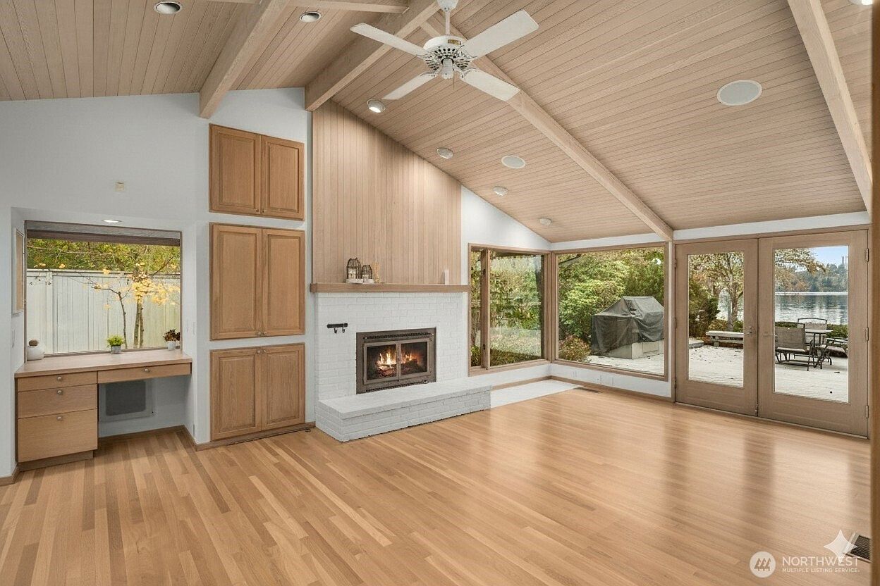 This spacious living room features a vaulted wood-paneled ceiling and light-toned hardwood flooring, creating a warm and airy atmosphere. A white brick fireplace with a wood mantel serves as a focal point, flanked by built-in light wood cabinetry and a desk area. Large windows and glass doors provide expansive views of the outdoor landscape and water, filling the room with natural light.