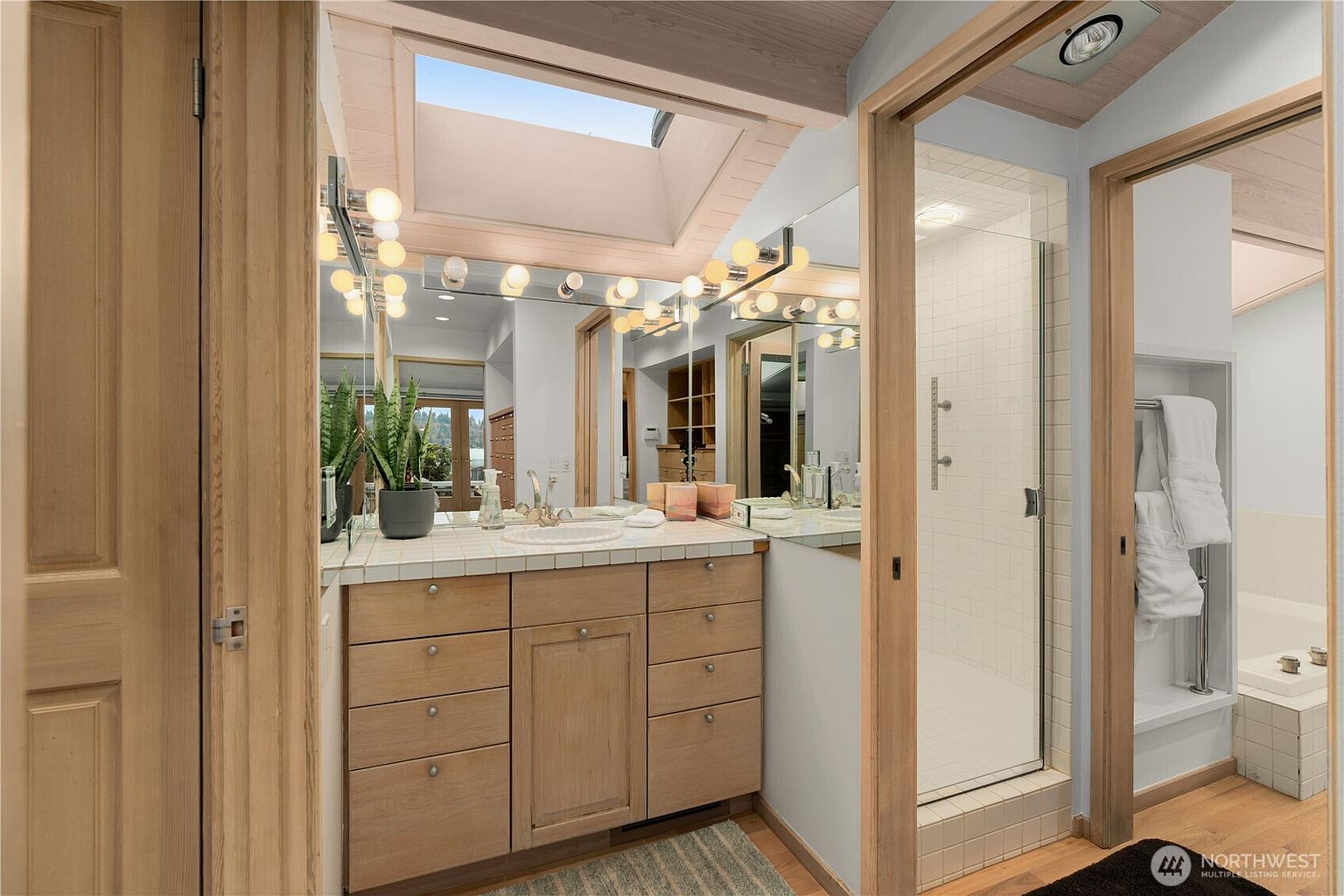 This primary bathroom features a light wood vanity with a tiled countertop and a large mirror illuminated by a series of globe lights. A skylight above provides natural light, while a glass-enclosed shower with white tile walls sits to the right. The space is framed by light wood door casings, creating a cohesive and bright aesthetic.