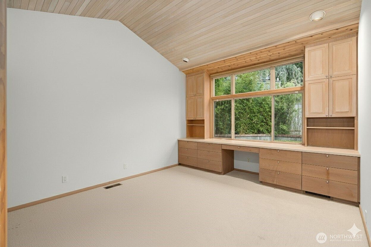 This bright home office features a vaulted wood-paneled ceiling and neutral carpeting, creating a warm and inviting workspace. A large built-in desk unit with light wood cabinetry and shelving spans the wall beneath a wide window, providing ample storage and a view of the outdoors. The room is clean and minimalist, offering a functional layout suitable for a dedicated study or home office.