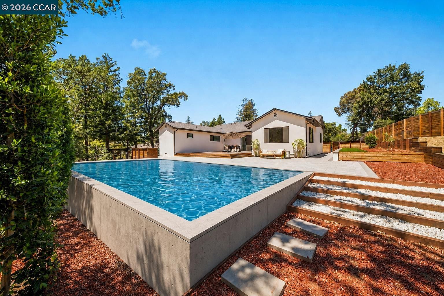 This backyard features a prominent, elevated rectangular swimming pool with a clean concrete surround, set against a backdrop of mature trees and a clear blue sky. A paved patio area extends from the rear of the light-colored house, offering space for outdoor dining and relaxation. The landscape includes tiered wooden retaining walls, gravel-filled steps, and mulch beds, creating a structured and modern outdoor living environment.