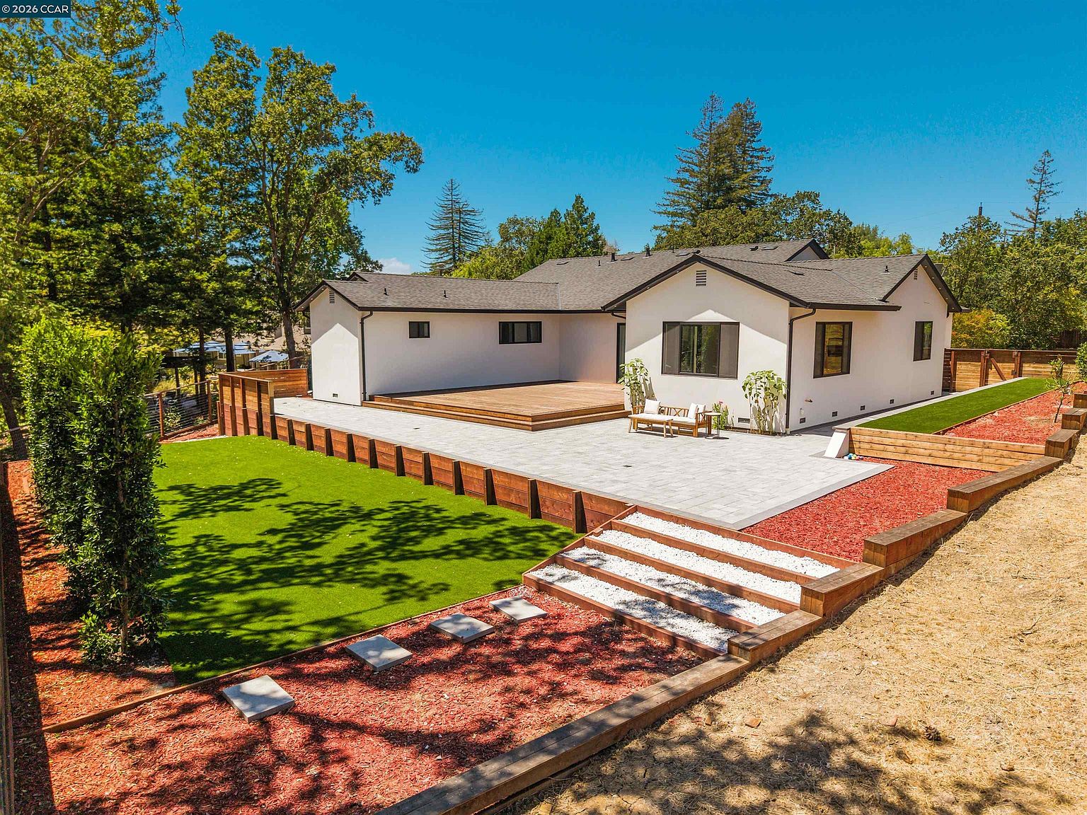 This elevated perspective captures the expansive rear exterior of a modern home, featuring a clean white facade with dark trim and large windows. The backyard is thoughtfully landscaped with a spacious light-toned paver patio, a raised wooden deck, and a vibrant green lawn bordered by tiered wooden retaining walls. A set of wide, gravel-filled steps leads down to a lower area covered in red mulch, creating a structured and inviting outdoor living space.