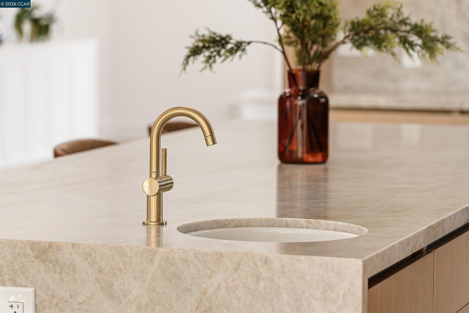 This close-up shot highlights a modern, gold-toned faucet mounted on a light-colored stone countertop with a subtle, integrated sink basin. A decorative amber glass vase filled with greenery sits in the background, adding a touch of organic warmth to the sleek, minimalist aesthetic. The perspective is eye-level, focusing on the clean lines and high-end finishes of the kitchen island fixture.