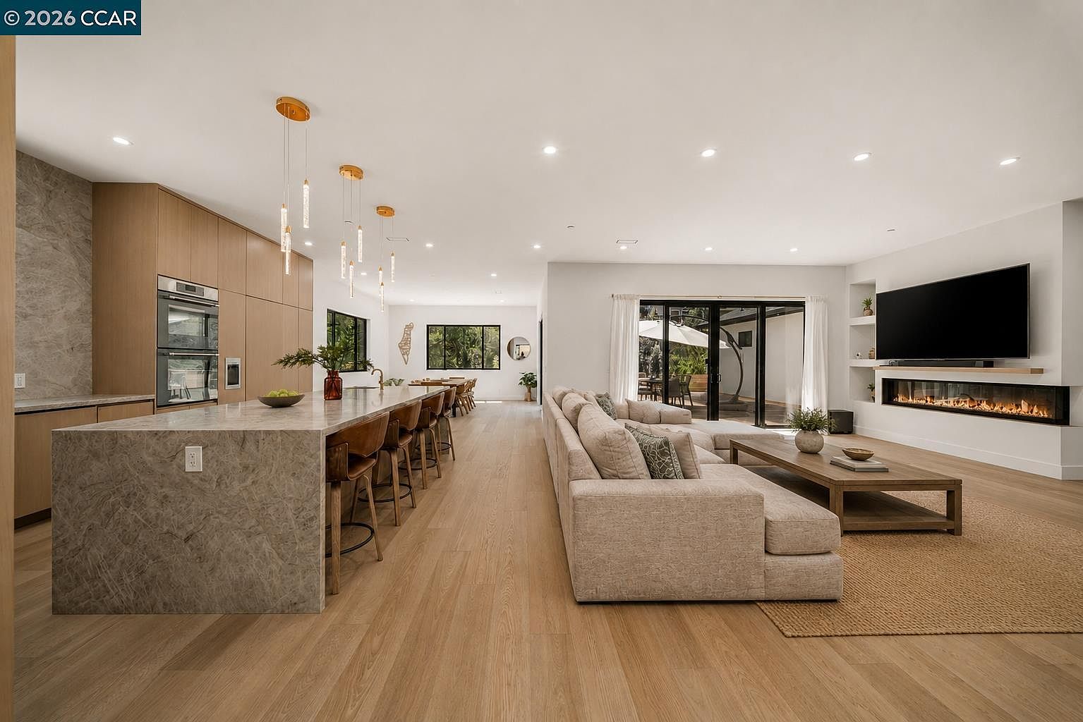This open-concept living space features a large kitchen island with a stone countertop and bar seating, seamlessly flowing into a spacious living area. The room is anchored by a linear fireplace set into a white wall with a television mounted above, while light wood flooring runs throughout the entire space. Large glass sliding doors at the rear provide a view of the outdoor patio, creating a bright and airy atmosphere perfect for modern entertaining.