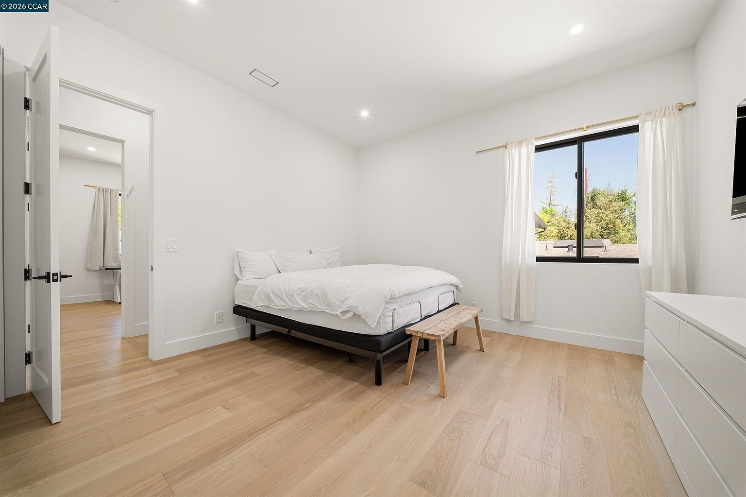 This bright and minimalist bedroom features light wood flooring and crisp white walls, creating an airy and clean atmosphere. A bed with white bedding is positioned against the wall, accompanied by a simple wooden bench at the foot. A window with white curtains allows natural light to fill the space, while a doorway leads to an adjacent room, showcasing the home's open and connected layout.