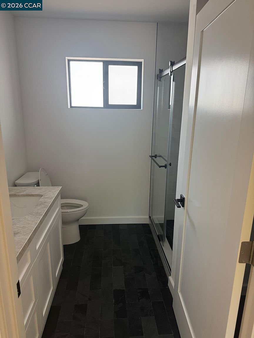 This compact, modern bathroom features a white vanity with a stone-look countertop and a dark, textured tile floor. To the right, a glass-enclosed shower with a sliding door adds a sleek, contemporary touch to the space. A window above the toilet provides natural light, creating a clean and functional atmosphere.