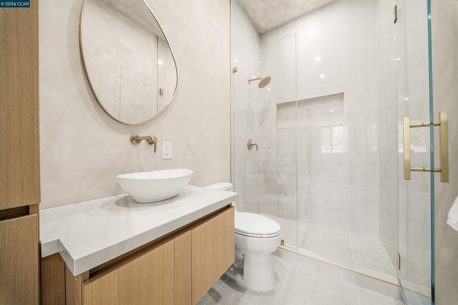 This modern bathroom features a light wood vanity topped with a white vessel sink and a wall-mounted faucet. A circular mirror hangs above the vanity, while a glass-enclosed shower with gold-toned fixtures and a recessed niche sits to the right. The space is finished with neutral, light-colored wall and floor tiles, creating a clean and contemporary aesthetic.
