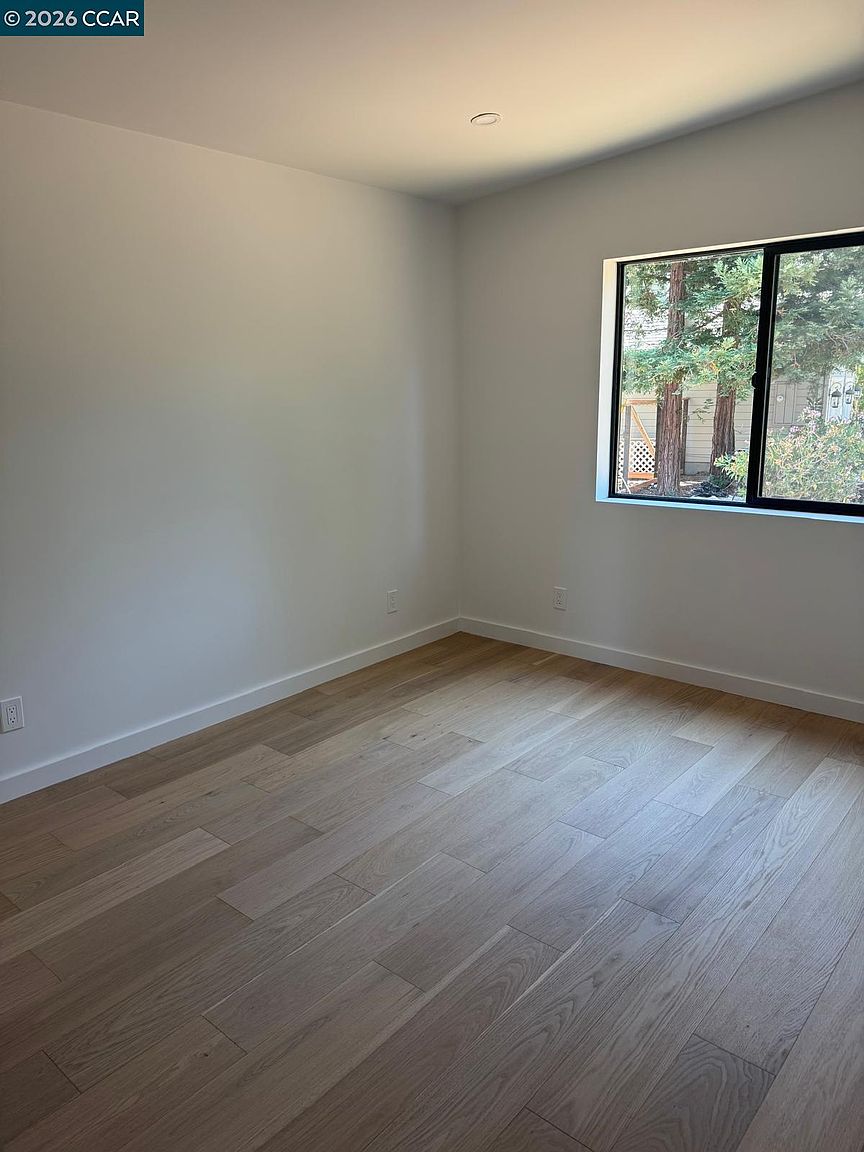 This bright, minimalist bedroom features light-toned wood-look flooring and neutral, off-white walls. A large window on the right wall provides natural light and a view of trees outside, while a single recessed light fixture is centered on the ceiling. The room is presented as a clean, blank canvas, offering a versatile space ready for personalization.