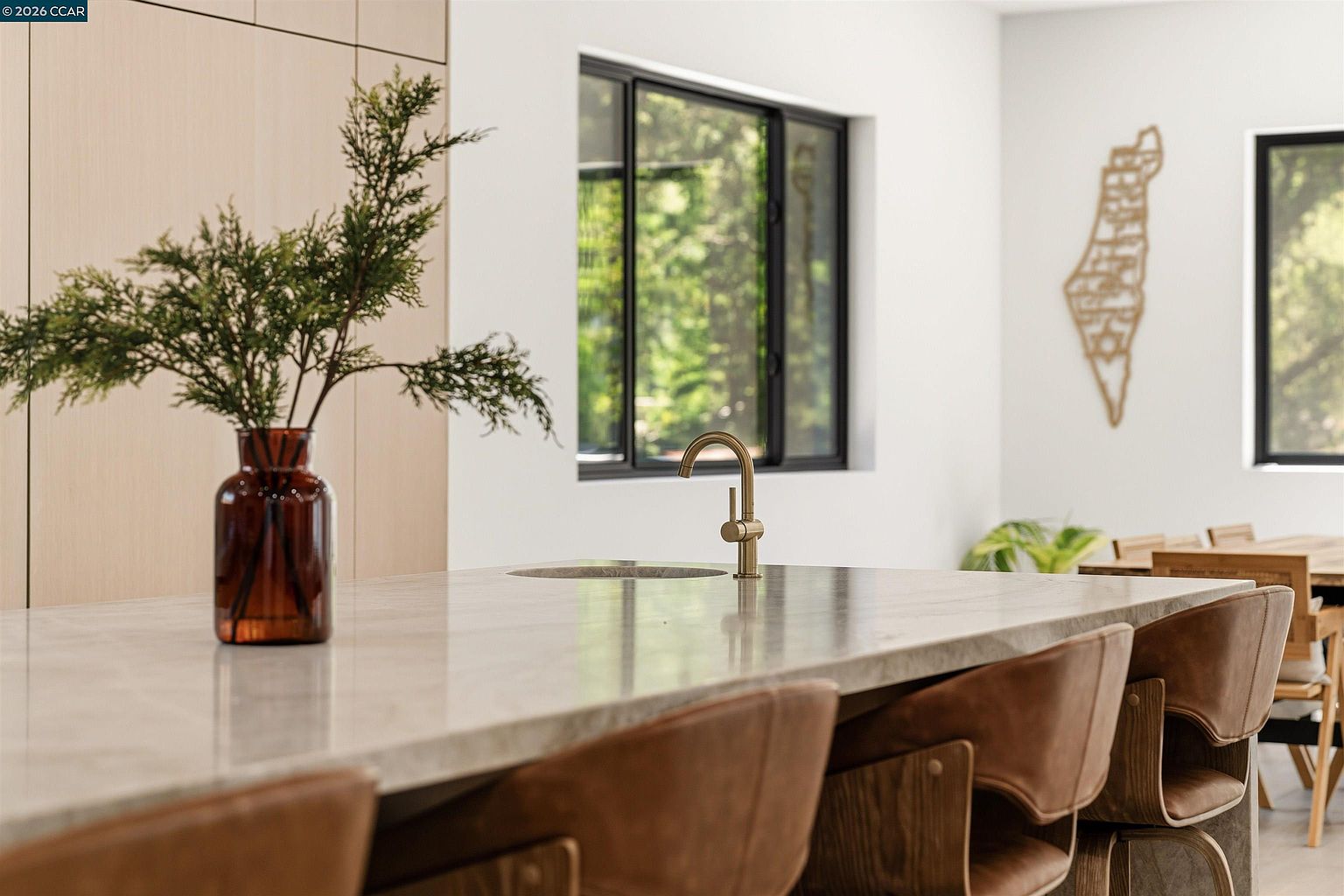 This modern kitchen features a large island with a stone countertop, a brass-toned faucet, and a circular sink. The space is accented by brown leather bar stools with light wood frames and a decorative vase with greenery, while a large window in the background provides natural light. The overall aesthetic is clean and contemporary, with light wood cabinetry visible in the background and a decorative wall art piece mounted on the white wall.