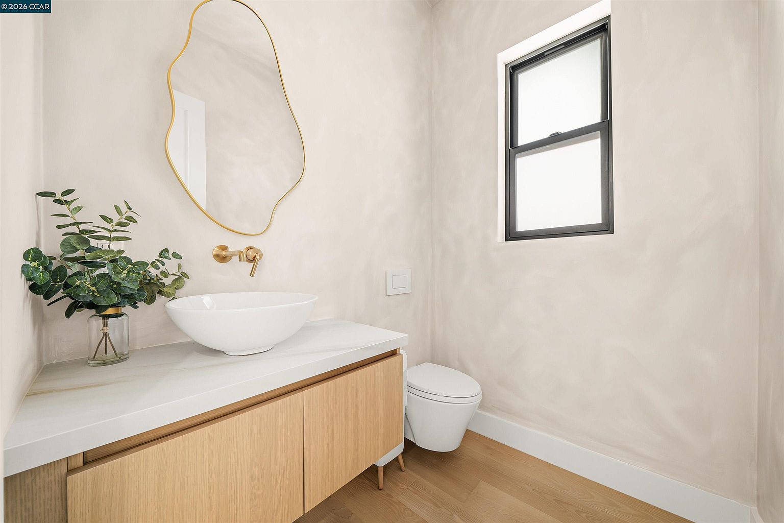This modern powder room features a minimalist aesthetic with light wood cabinetry, a white vessel sink, and a unique organic-shaped mirror with a gold frame. The walls are finished in a soft, textured neutral tone, complemented by light wood flooring and a wall-mounted toilet. A window provides natural light, while a decorative plant adds a touch of greenery to the vanity area.