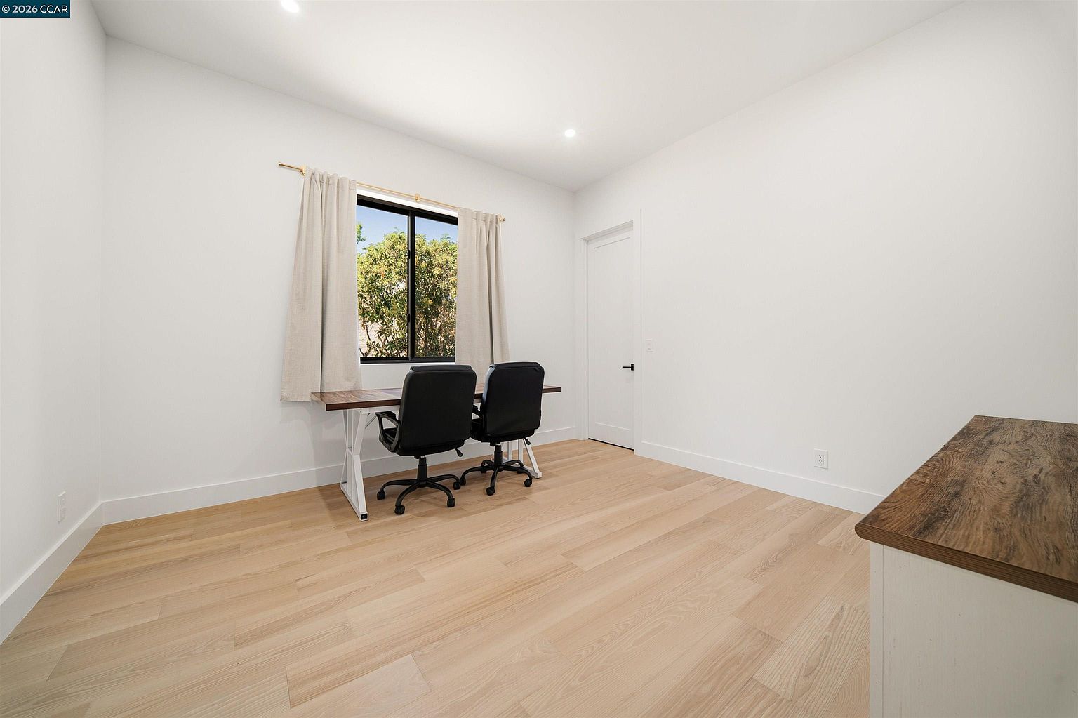 This bright and minimalist home office features light wood-toned flooring and crisp white walls, creating an airy and productive atmosphere. A long desk positioned under a window provides a dedicated workspace, complemented by two black office chairs. The room is simple and uncluttered, offering a versatile space that could easily serve as a study or remote work area.