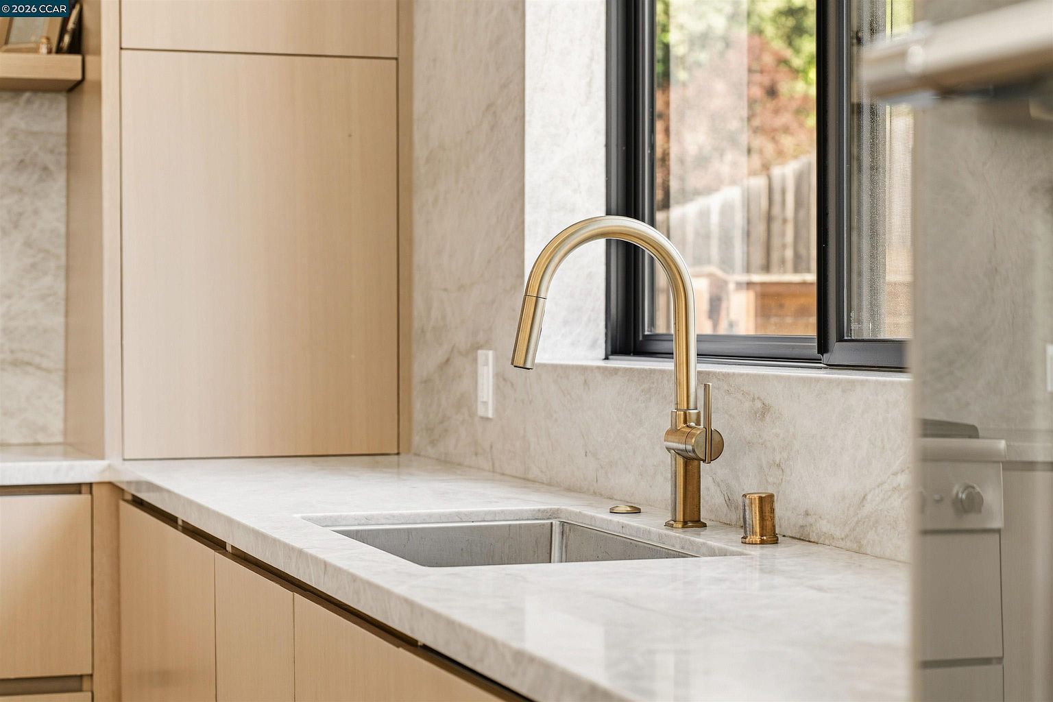 This close-up shot highlights a modern kitchen sink area featuring a sleek, brass-toned gooseneck faucet and a matching soap dispenser. The countertop and backsplash are finished in a light-toned stone with subtle veining, complemented by light wood cabinetry. A window with a dark frame sits behind the sink, providing a view of the outdoors and adding natural light to the workspace.