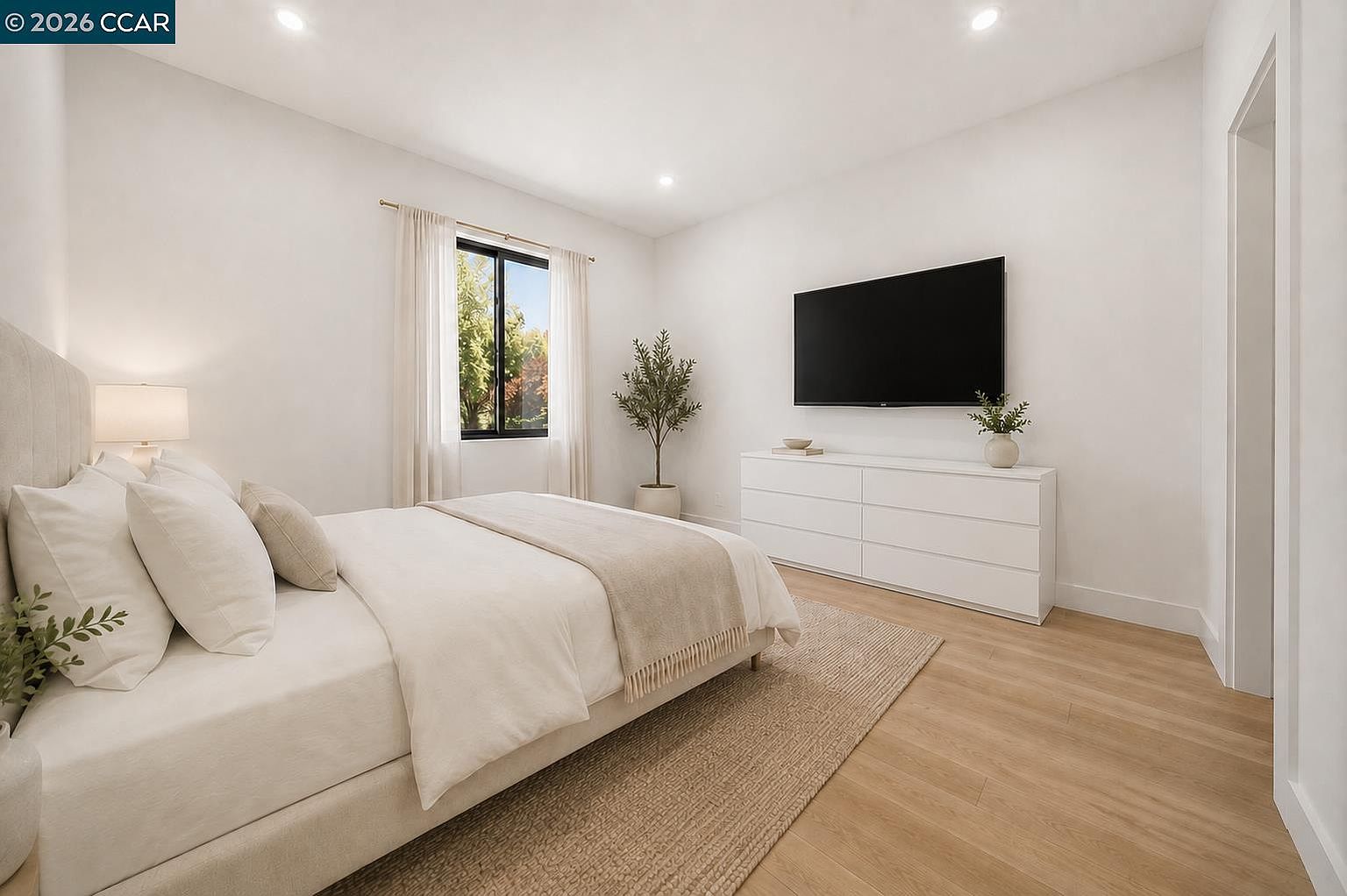 This bright and airy bedroom features a neutral color palette with light wood-toned flooring and crisp white walls. A plush bed with layered linens sits on a textured area rug, while a sleek white dresser positioned beneath a wall-mounted television provides clean storage. The room is accented by a potted plant and sheer curtains that frame a window, creating a serene and inviting atmosphere.