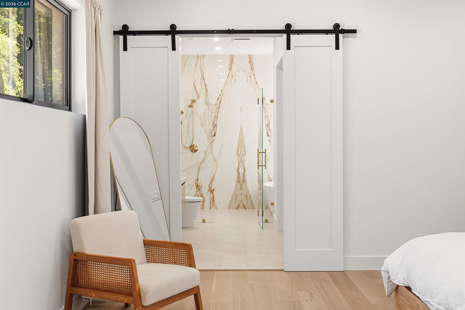 This view from a bedroom highlights a modern primary bathroom accessed through double white barn doors. The bathroom features a striking, large-format patterned stone wall behind a glass-enclosed shower and a freestanding tub. In the foreground, a light wood-framed accent chair with woven detailing sits next to an oval floor mirror, creating a sophisticated and airy atmosphere.