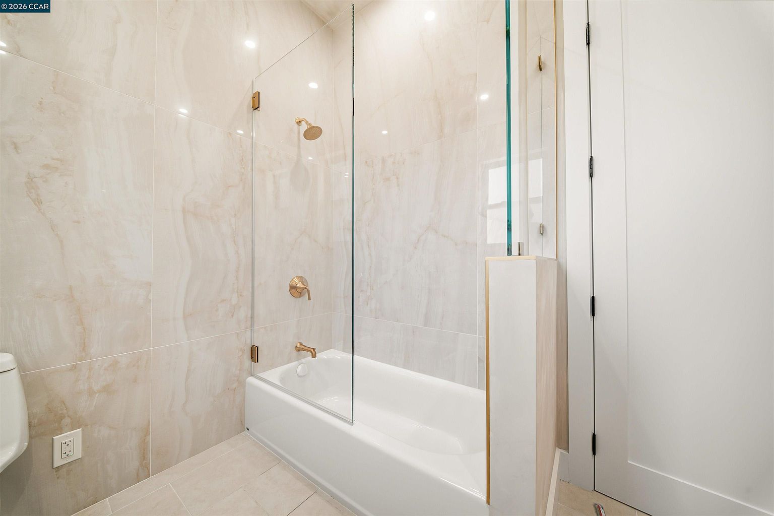 This modern bathroom features a shower-tub combination enclosed by a frameless glass panel. The walls are finished with large, light-toned tiles with subtle veining, complemented by gold-toned plumbing fixtures. A partial wall with a gold-trimmed edge separates the tub area from the rest of the room, creating a clean and contemporary aesthetic.