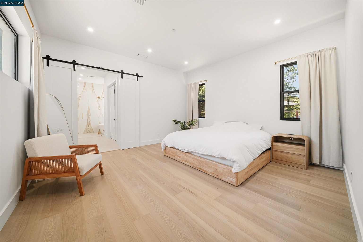 This bright and airy primary bedroom features light wood flooring and a minimalist aesthetic with white walls. A low-profile light wood bed frame is centered in the room, complemented by a matching curved nightstand and a modern accent chair with cane detailing. A sliding barn door provides access to an adjacent bathroom, while large windows with neutral curtains allow for ample natural light.