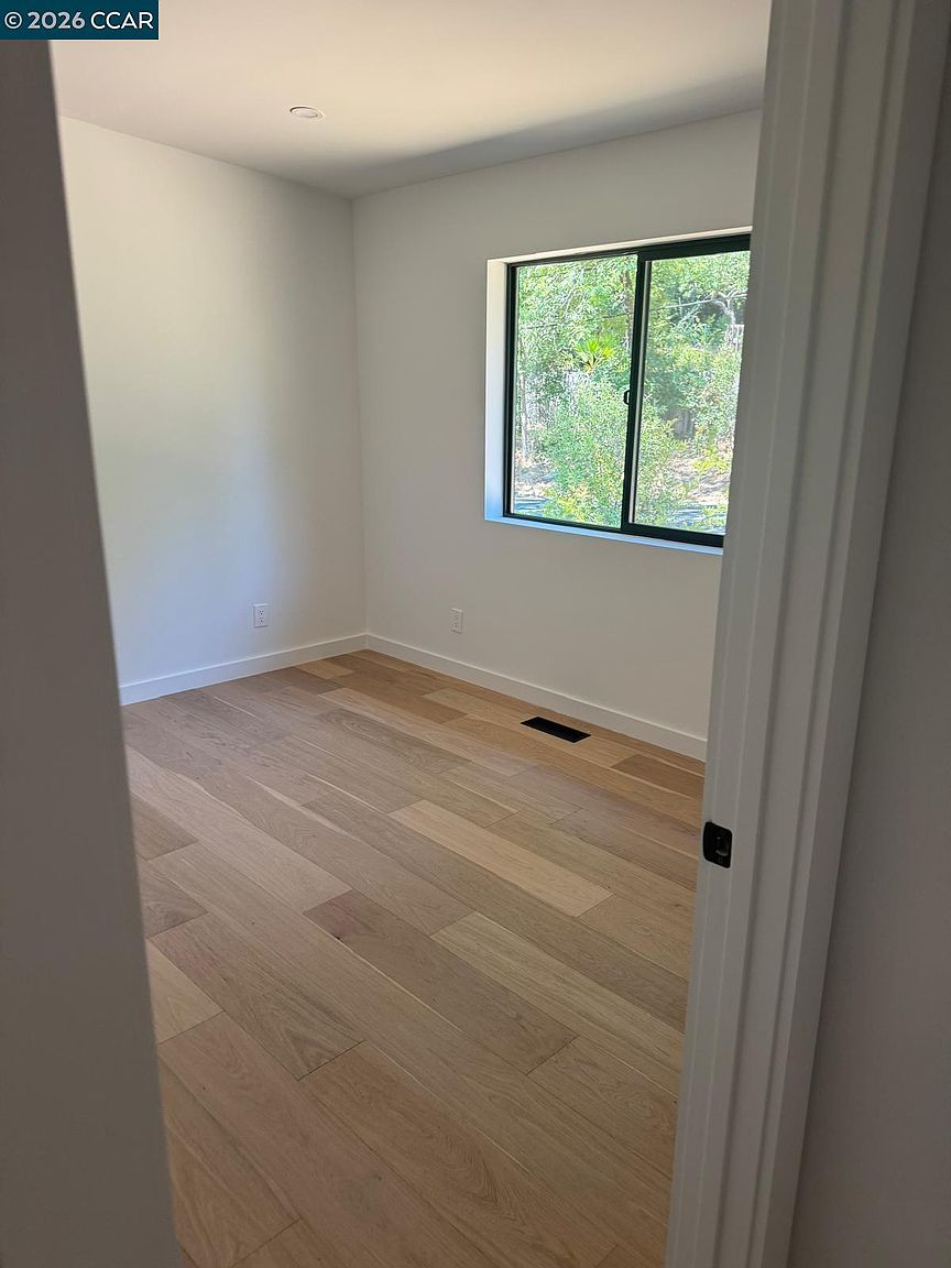 This image shows a bright, unfurnished bedroom viewed from the doorway, highlighting light-toned wood flooring and neutral-colored walls. A single window on the far wall provides natural light and a view of the surrounding greenery. The space is clean and minimalist, offering a versatile canvas for potential residents.
