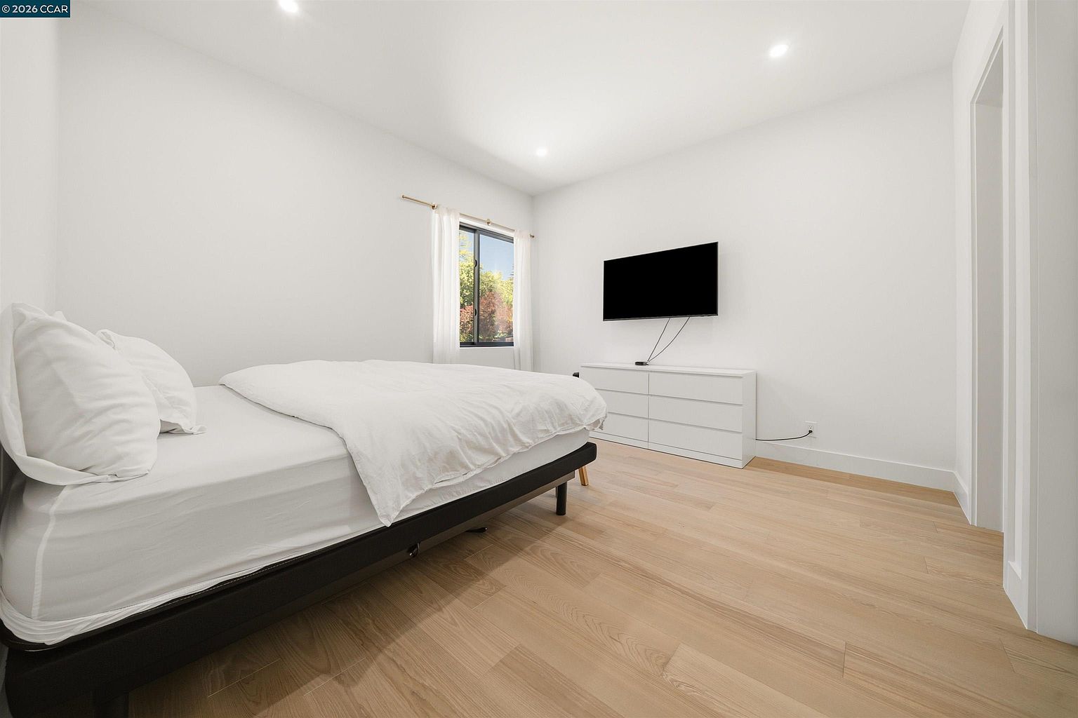 This minimalist bedroom features light wood-toned flooring and crisp white walls, creating a bright and airy atmosphere. A bed with white bedding is positioned on the left, while a white dresser sits against the wall beneath a wall-mounted television. A single window with white curtains allows natural light to enter, providing a clean and simple aesthetic suitable for a guest room or secondary bedroom.
