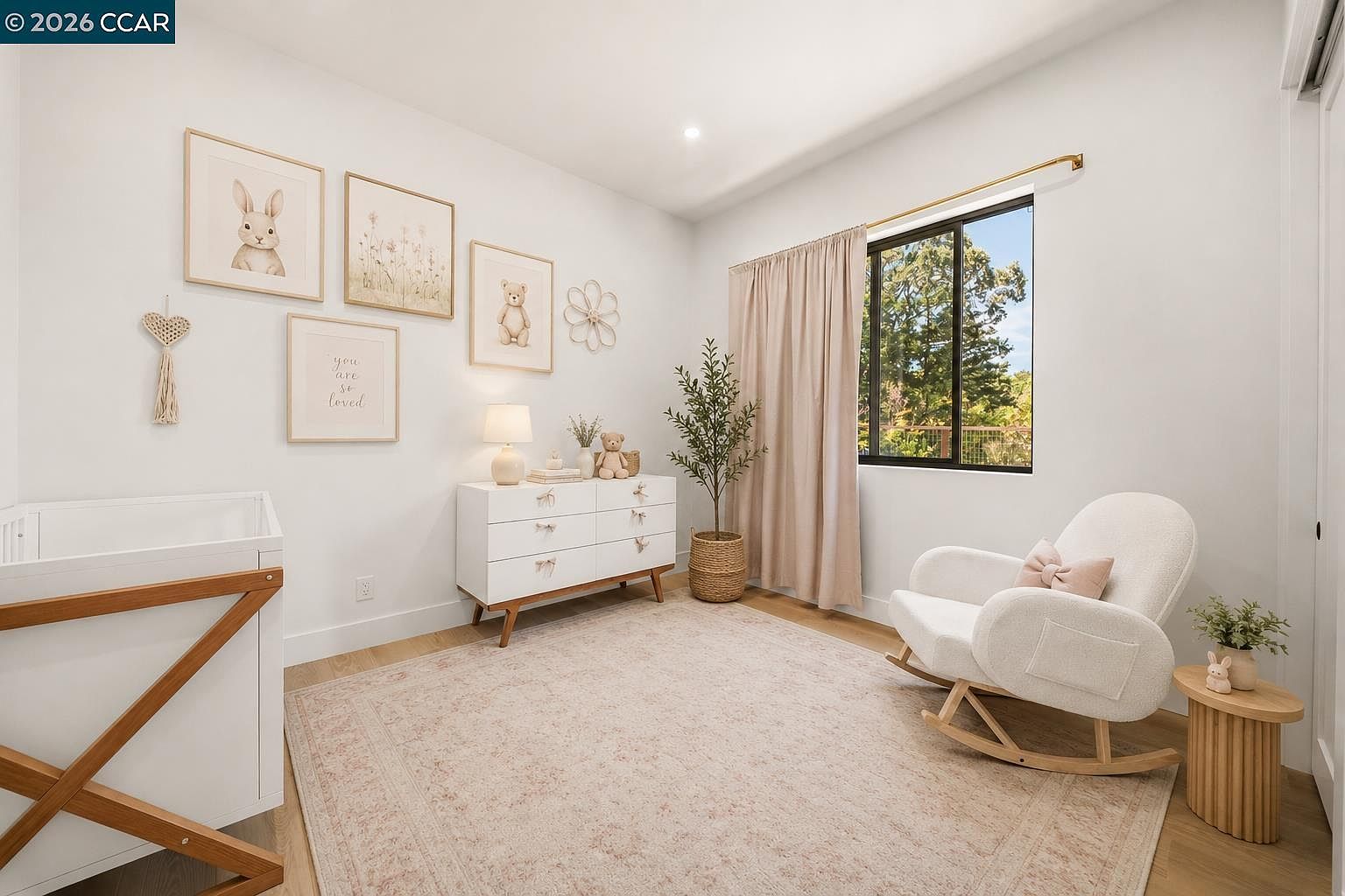 This charming nursery features a soft, neutral color palette with light wood flooring and a large area rug. The room is furnished with a white dresser, a textured rocking chair, and a crib, accented by whimsical wall art and a potted plant. A window with light-colored curtains allows natural light to fill the space, creating a warm and inviting atmosphere.