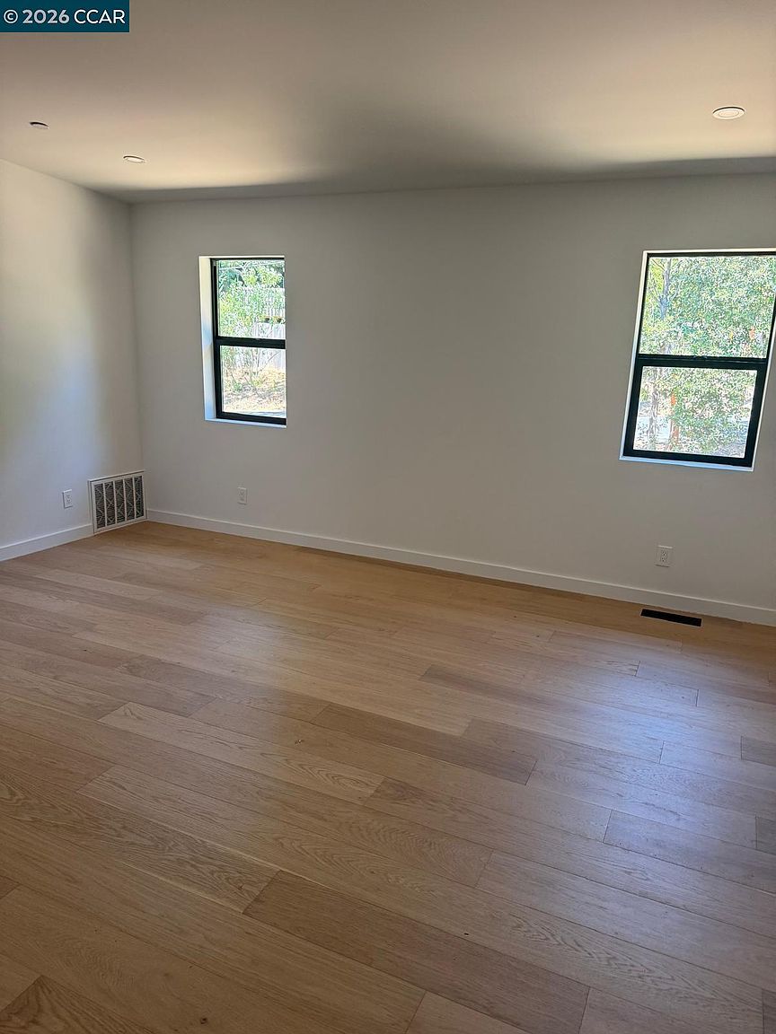 This bright, unfurnished room features light-toned wood flooring and neutral white walls, creating a clean and versatile living space. Two windows allow for natural light to fill the room, while a wall-mounted vent and a floor-level vent are visible. The perspective is from the doorway, looking across the open floor plan toward the exterior-facing walls.
