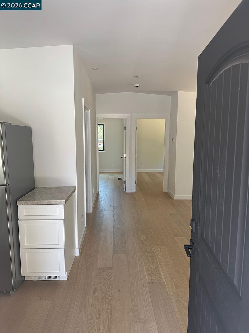This interior view captures a bright hallway leading from a kitchen area toward two separate doorways. The space features light-toned wood flooring and crisp white walls, creating a clean and modern aesthetic. A small cabinet with a stone-look countertop sits adjacent to a stainless steel refrigerator on the left, while a dark-colored door frame is visible in the foreground.