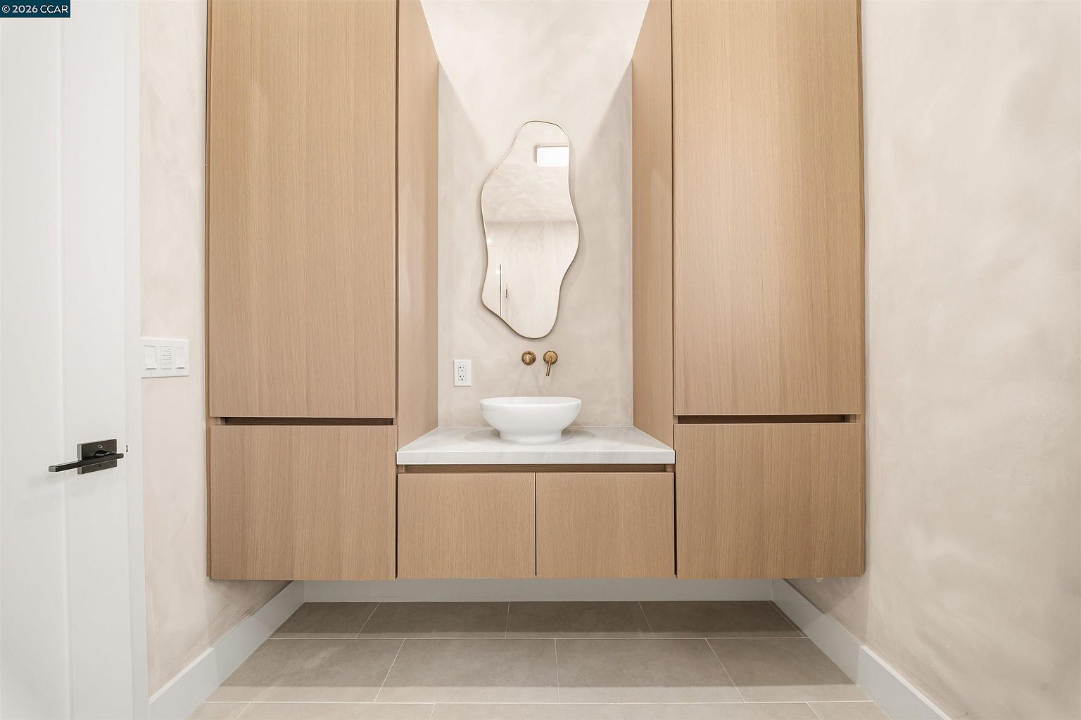 This minimalist powder room features a floating vanity with light wood cabinetry and a white vessel sink set against a neutral, textured wall. An organic-shaped mirror hangs above the sink, flanked by tall, integrated wood storage cabinets that provide a clean, symmetrical aesthetic. The space is finished with large-format light-toned floor tiles, creating a serene and contemporary atmosphere.