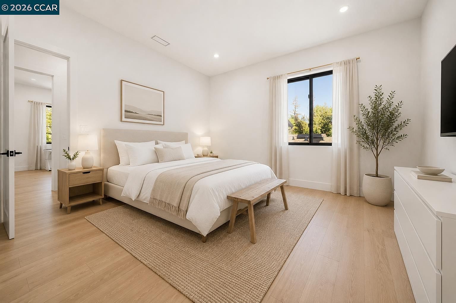 This bright and airy primary bedroom features light wood-toned flooring and a neutral color palette that creates a serene atmosphere. A plush bed with a light-colored upholstered headboard serves as the focal point, flanked by matching nightstands and lamps, while a wooden bench sits at the foot of the bed atop a textured area rug. Large windows draped in soft curtains allow natural light to fill the space, and a white dresser provides additional storage, all captured from a wide-angle perspective that emphasizes the room's spaciousness.