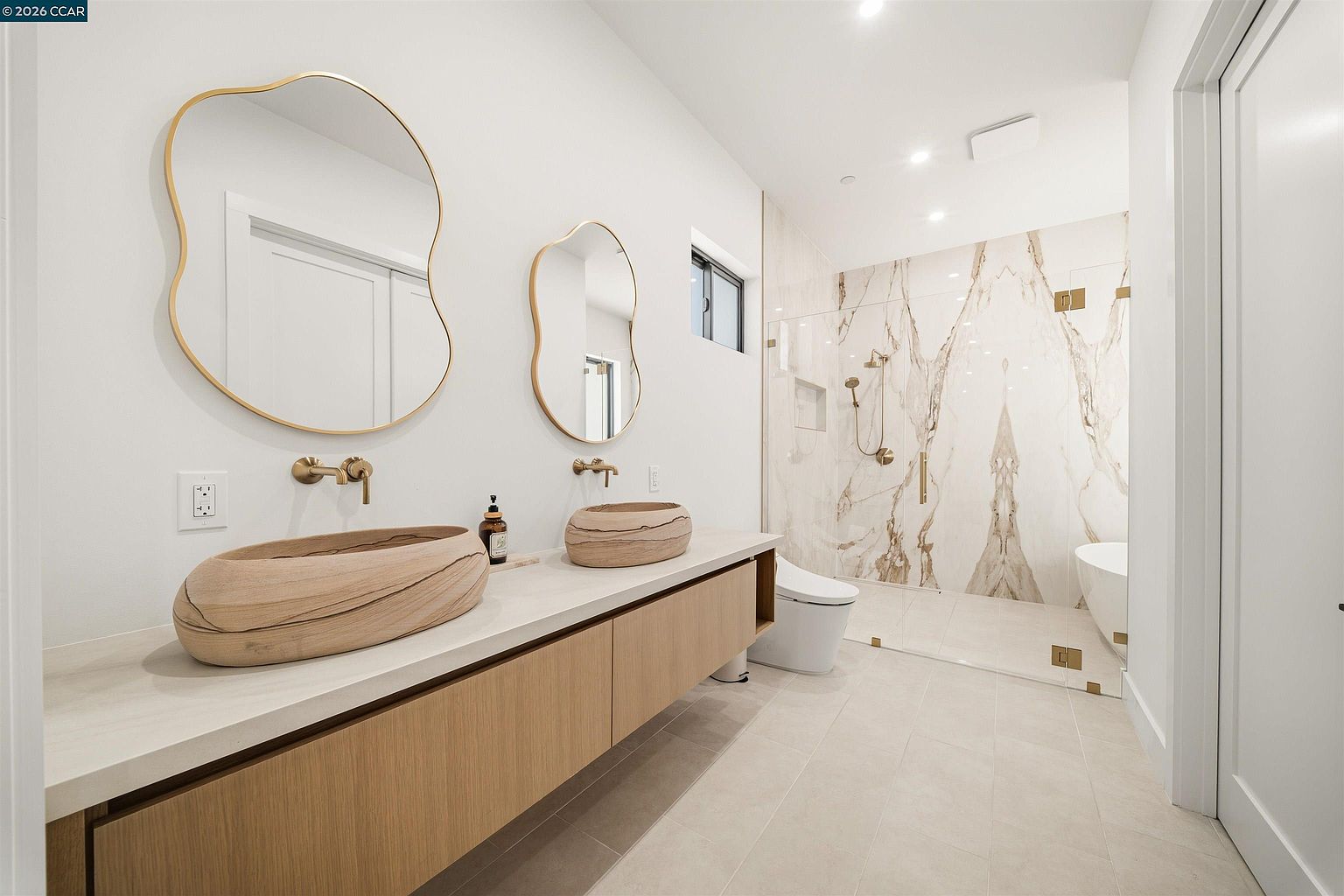 This modern primary bathroom features a dual-vanity with light wood cabinetry and two unique, organic-shaped stone vessel sinks. Above the vanity, two asymmetrical mirrors with gold-toned frames are mounted on a clean white wall, complemented by wall-mounted gold fixtures. A glass-enclosed wet room area contains a shower with large-format patterned stone wall tiles and a separate soaking tub, creating a luxurious and spa-like atmosphere.