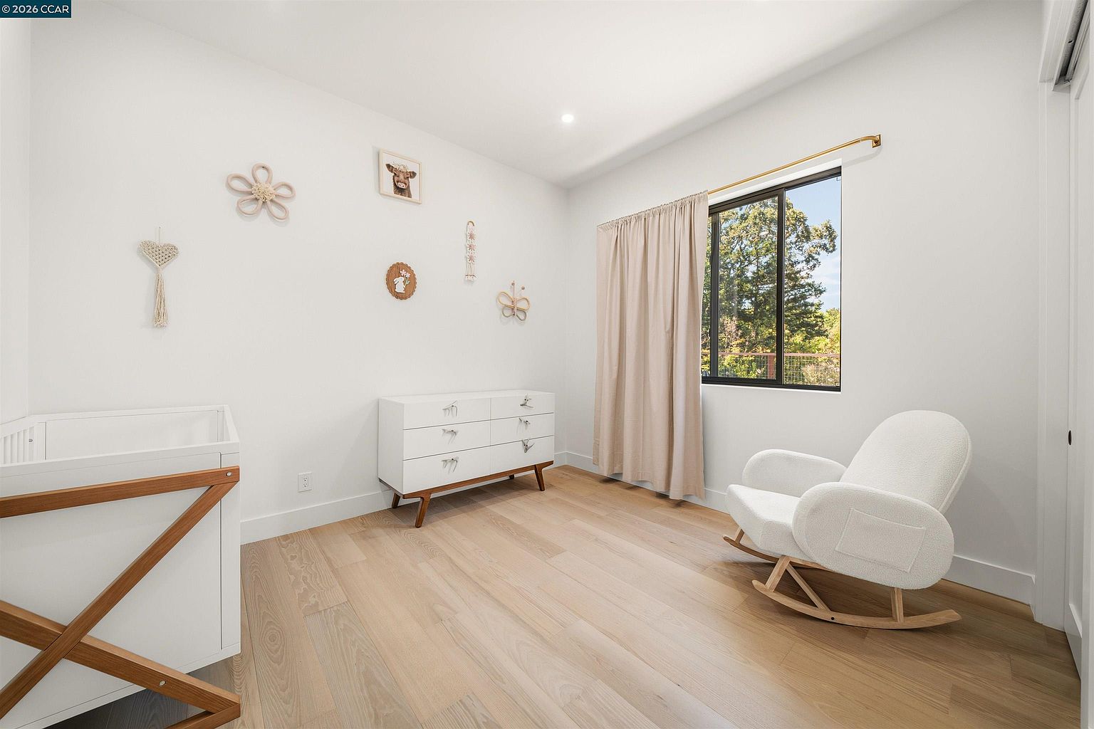 This bright and airy nursery features light wood flooring, crisp white walls, and a minimalist aesthetic. The room is furnished with a white dresser, a modern white rocking chair, and a crib, accented by delicate wall decor. A window with soft beige curtains allows natural light to fill the space, creating a calm and inviting atmosphere.