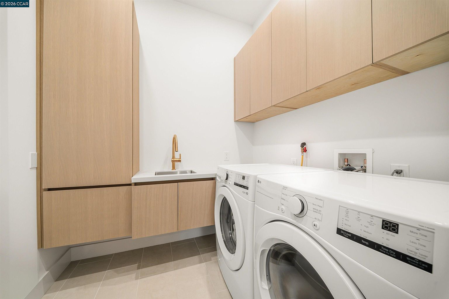 This modern laundry room features light wood cabinetry, including a tall storage unit and upper cabinets, paired with a clean white countertop and a gold-toned faucet. A side-by-side washer and dryer set is positioned beneath the counter, with neutral-toned floor tiles completing the minimalist aesthetic. The perspective is a wide-angle shot from the corner, showcasing the efficient layout and contemporary design of the utility space.
