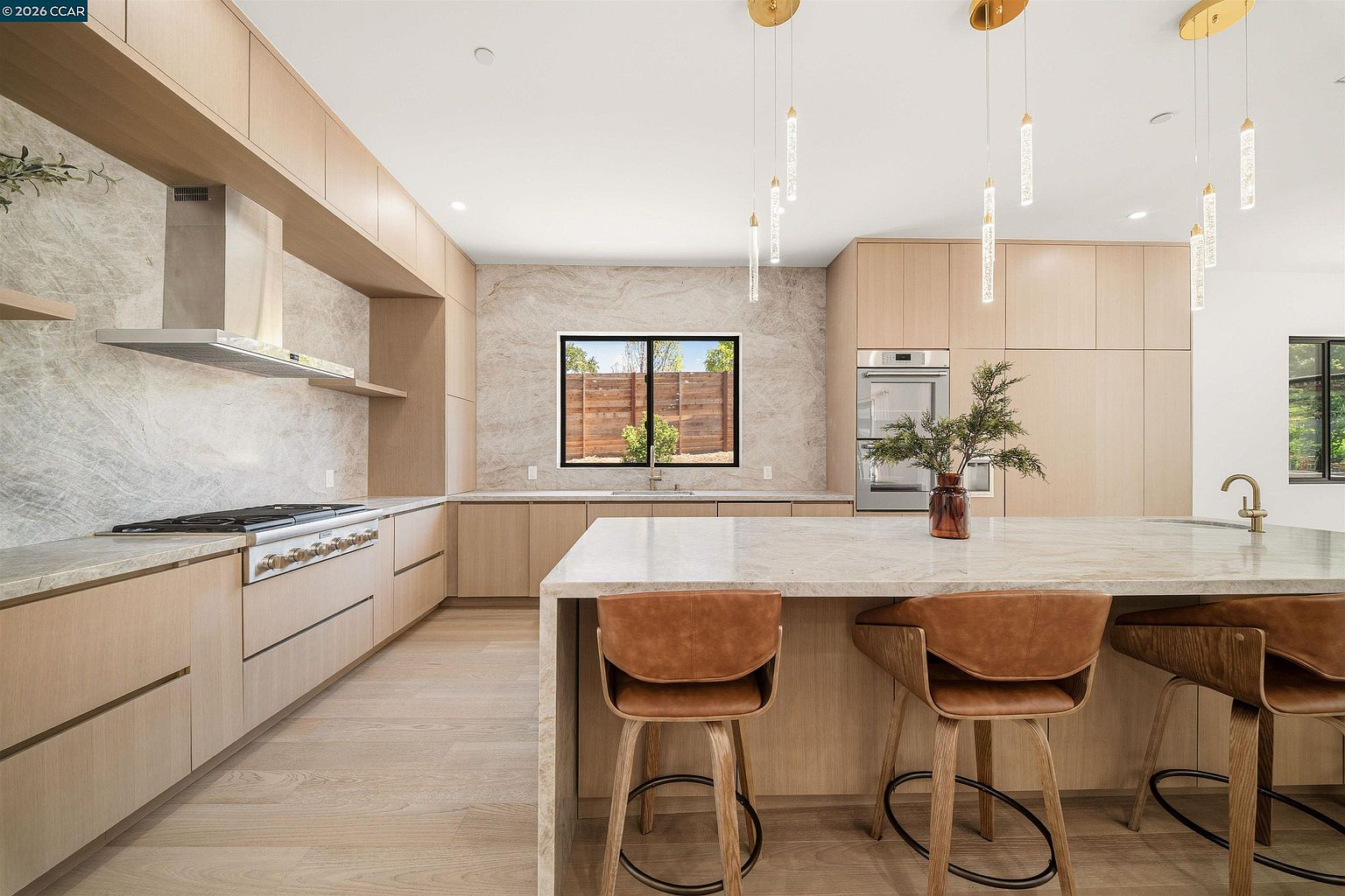 This modern, minimalist kitchen features light wood cabinetry and a large central island with stone countertops and leather-upholstered bar stools. A stone backsplash extends behind the range and sink area, complemented by sleek, integrated appliances and elegant pendant lighting. The perspective is a wide, eye-level shot that captures the clean lines and open layout of the space.