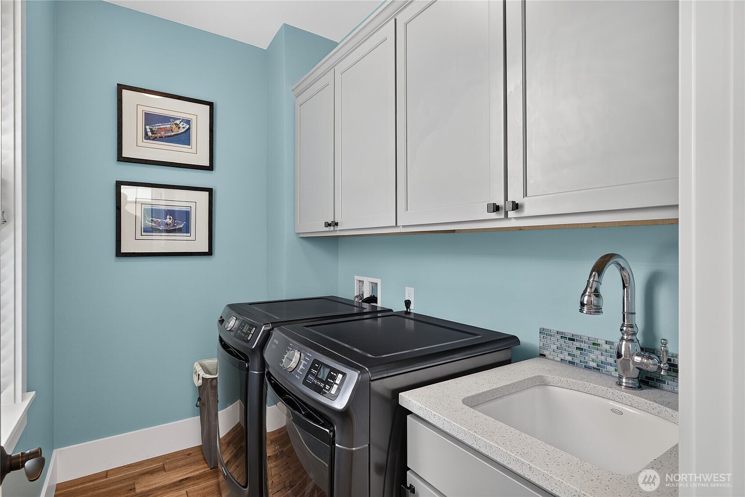 This functional laundry room features light blue walls, white upper cabinetry, and a sink with a stone countertop. A pair of dark-toned, modern laundry appliances are positioned beneath the cabinets, complemented by a small mosaic tile backsplash. The room is accented by two framed nautical-themed pictures on the wall and warm-toned wood flooring.