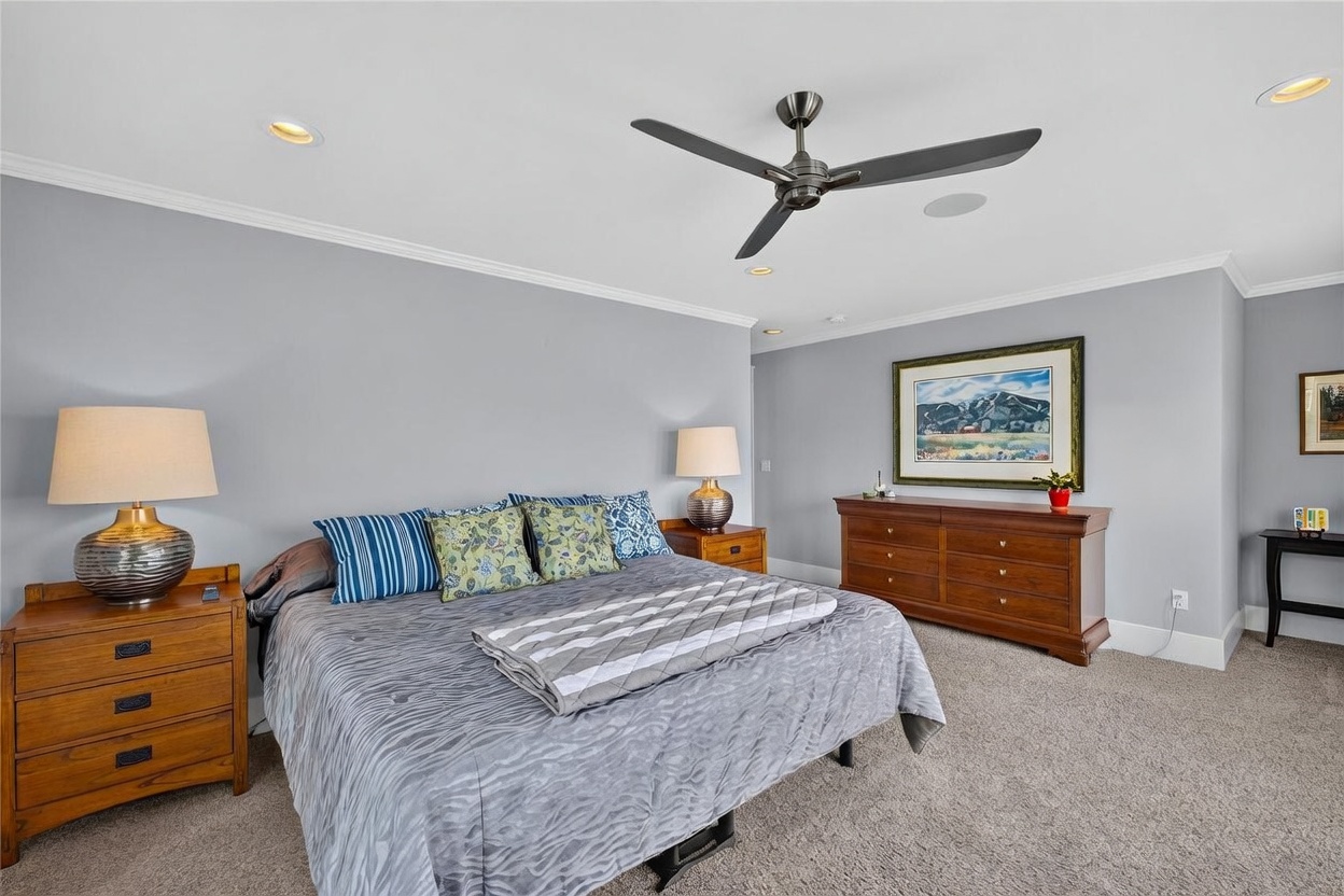 This spacious primary bedroom features neutral gray walls, plush carpeting, and a large bed centered against the wall with matching wooden nightstands and lamps. A wooden dresser sits against the opposite wall, topped with a framed landscape painting and a decorative floor lamp, creating a warm and inviting atmosphere. The room is illuminated by recessed lighting and a modern ceiling fan, offering a comfortable and well-appointed living space.