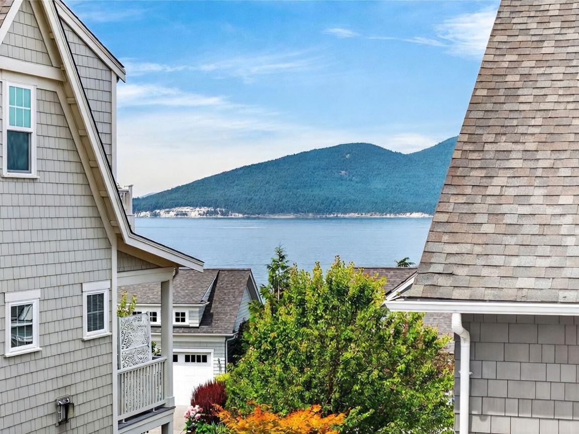 This view from an elevated balcony captures a stunning coastal landscape featuring a ferry crossing the water against a backdrop of forested mountains. The foreground is framed by the shingled siding of two neighboring homes, creating a sense of privacy and community. The scene emphasizes the property's scenic location and connection to the natural environment.
