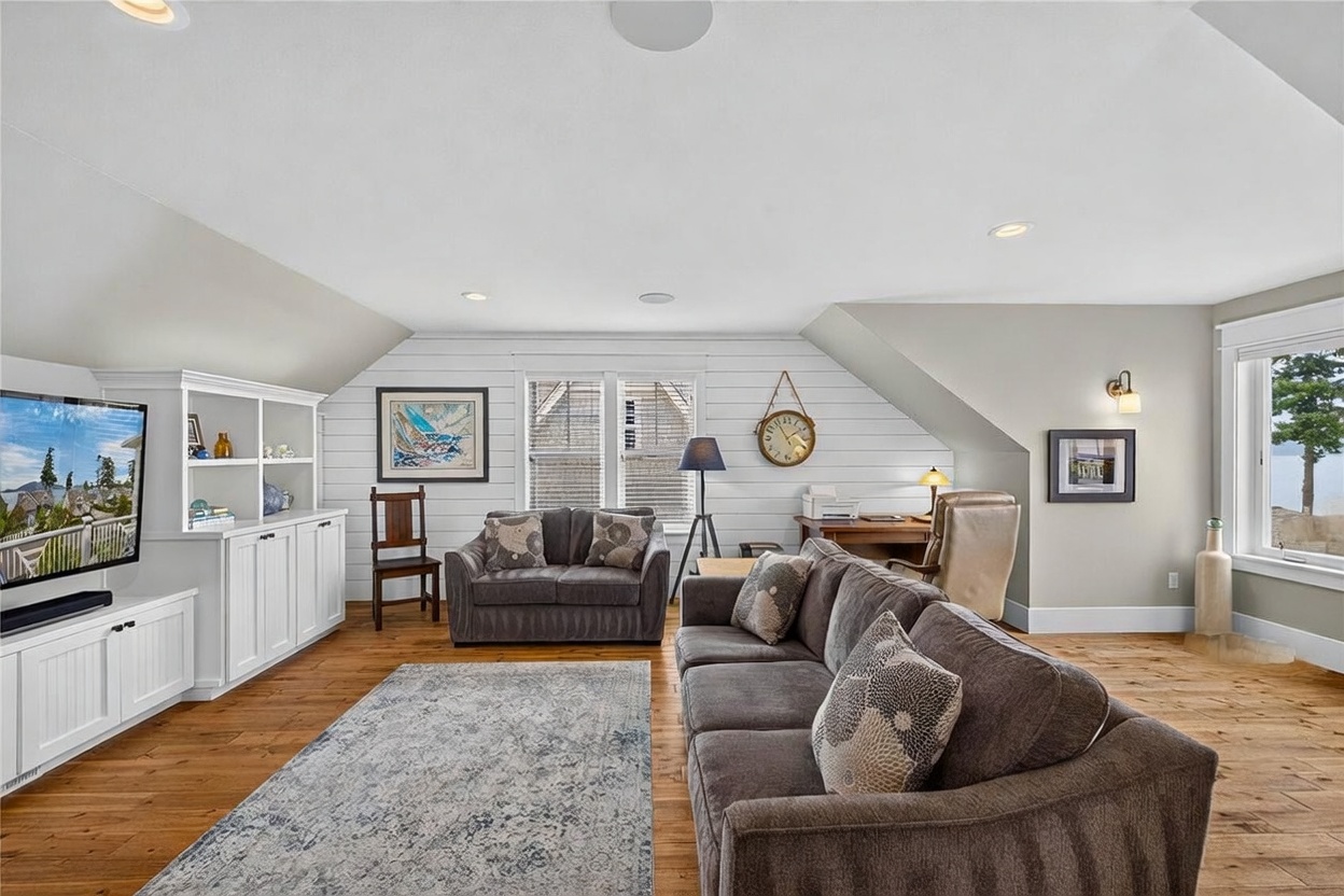 This spacious, light-filled living room features warm wood flooring and a shiplap accent wall that adds a coastal, relaxed aesthetic. The room is furnished with a large, comfortable grey sectional and a matching loveseat, complemented by a patterned area rug and a built-in white media console with shelving. A dedicated workspace with a desk and chair is tucked into the corner, while large windows provide ample natural light and views of the outdoors.