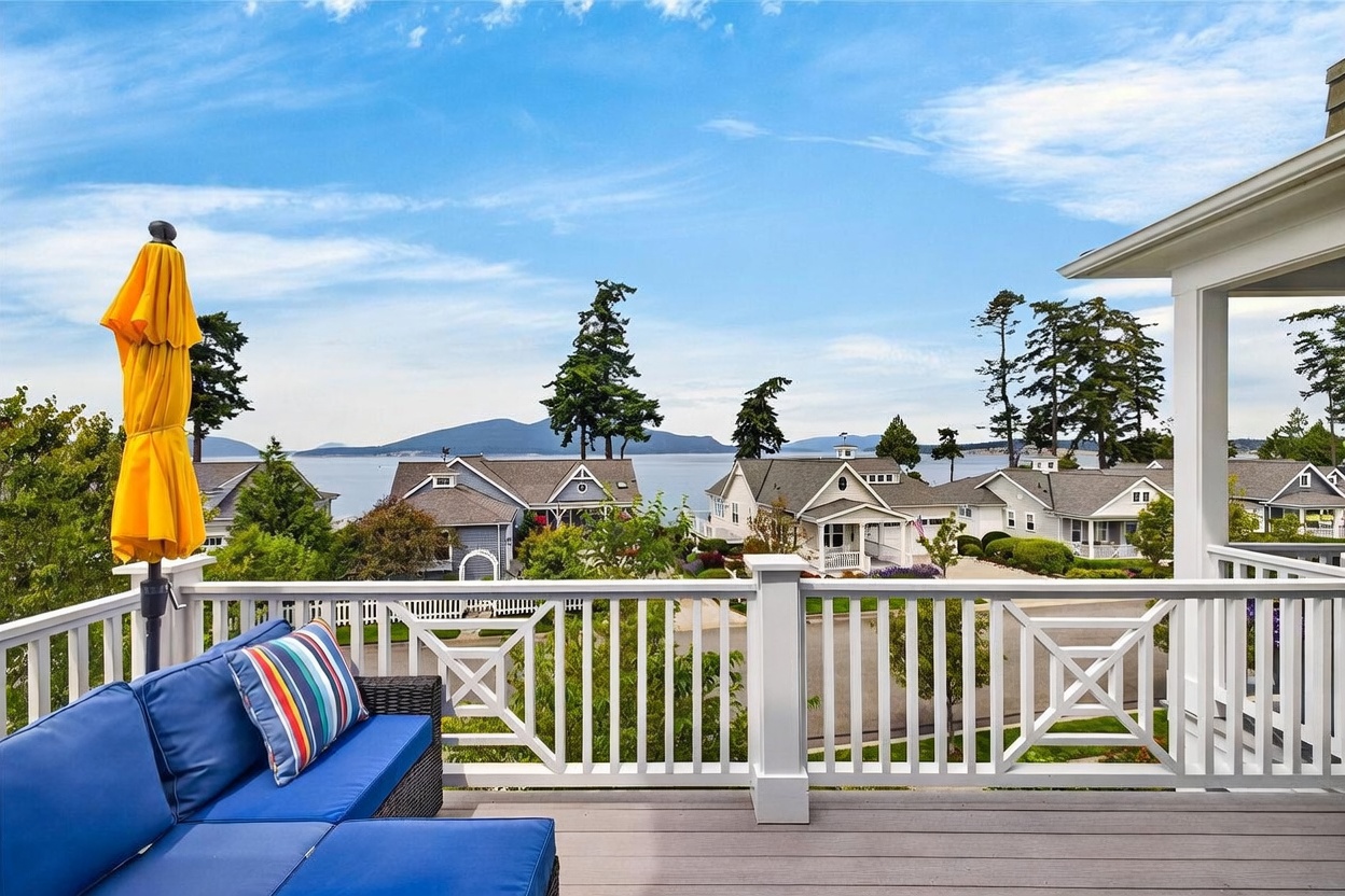 This elevated deck offers a stunning, expansive view of a calm bay surrounded by distant mountains and lush evergreen trees. The space is furnished with a comfortable blue sectional sofa featuring a colorful striped throw pillow and is shaded by a bright yellow umbrella. The white railing provides a clean, classic aesthetic, framing the picturesque neighborhood and waterfront scenery below.