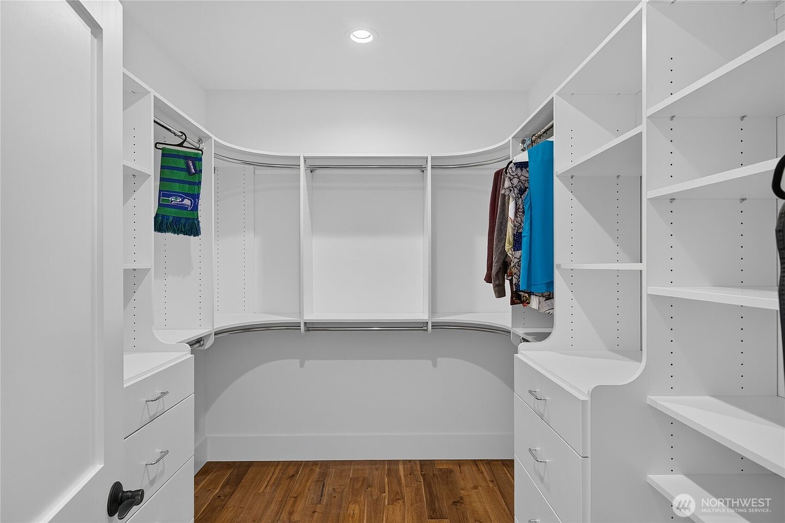 This walk-in closet features custom white built-in shelving and cabinetry with integrated hanging rods and drawers. The space is finished with warm-toned wood flooring and bright, recessed ceiling lighting. The symmetrical layout provides ample storage and organization, creating a clean and functional aesthetic.