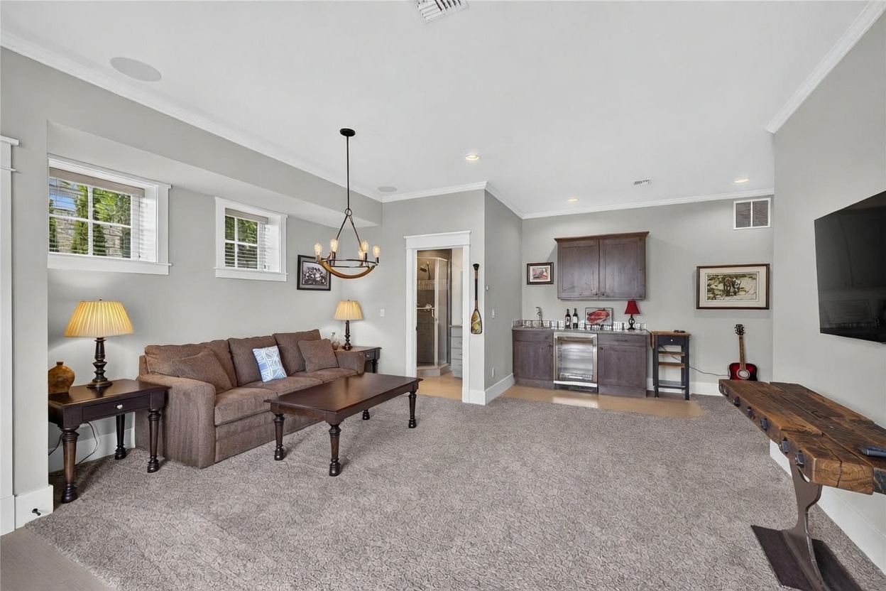 This spacious basement living area features neutral gray walls, plush carpeting, and a comfortable brown sofa arranged around a dark wood coffee table. A built-in wet bar with dark cabinetry, a beverage refrigerator, and a sink provides a convenient space for entertaining. The room is illuminated by a decorative chandelier and recessed lighting, with a doorway leading to an adjacent bathroom.