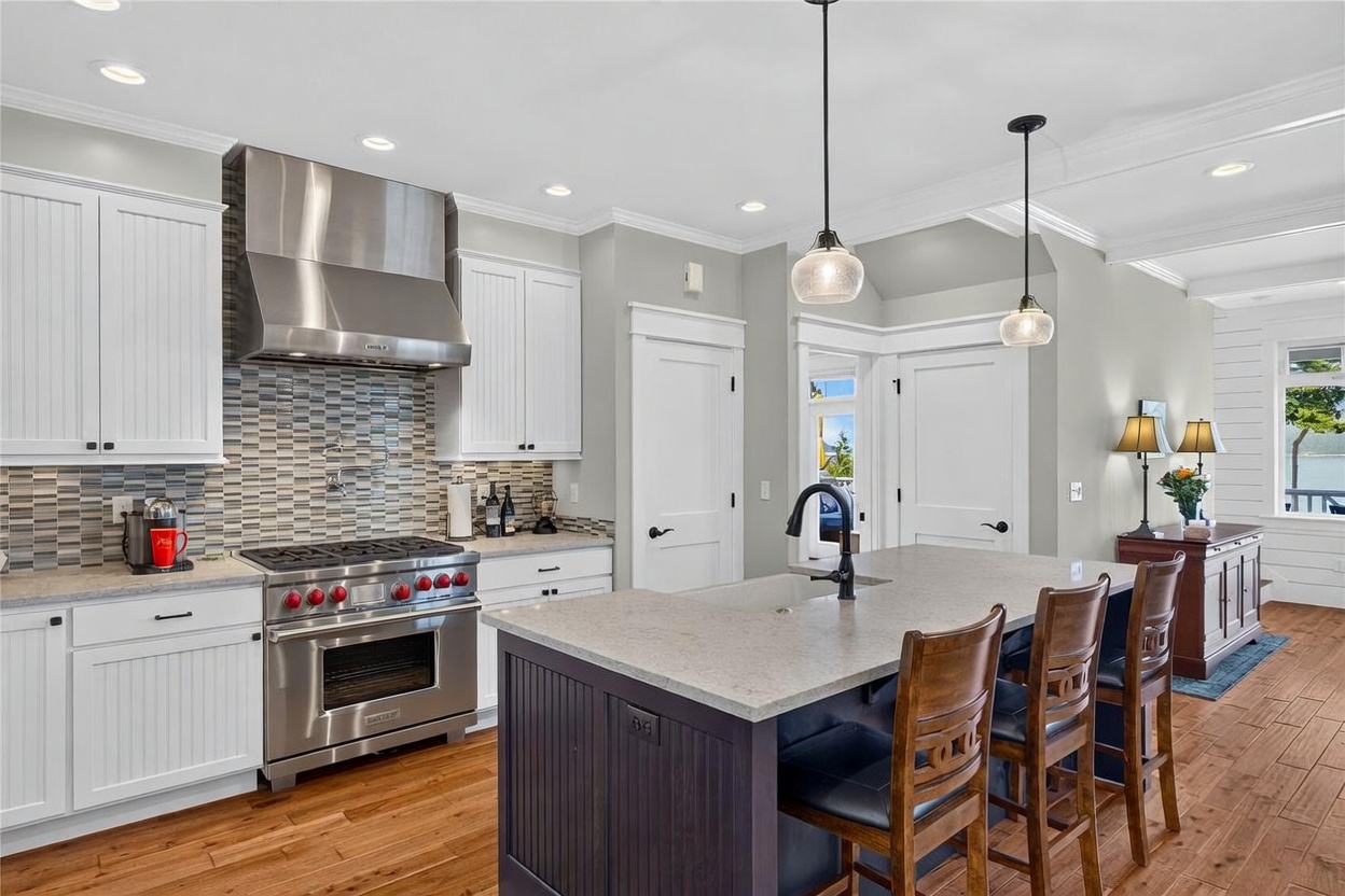 This bright and airy kitchen features white cabinetry paired with a dark-toned central island and light-colored stone countertops. A professional-grade stainless steel range with a matching hood is set against a decorative mosaic tile backsplash, while two pendant lights hang above the island seating. The space is finished with warm wood flooring and flows seamlessly into an adjacent living area with a view of the outdoors.