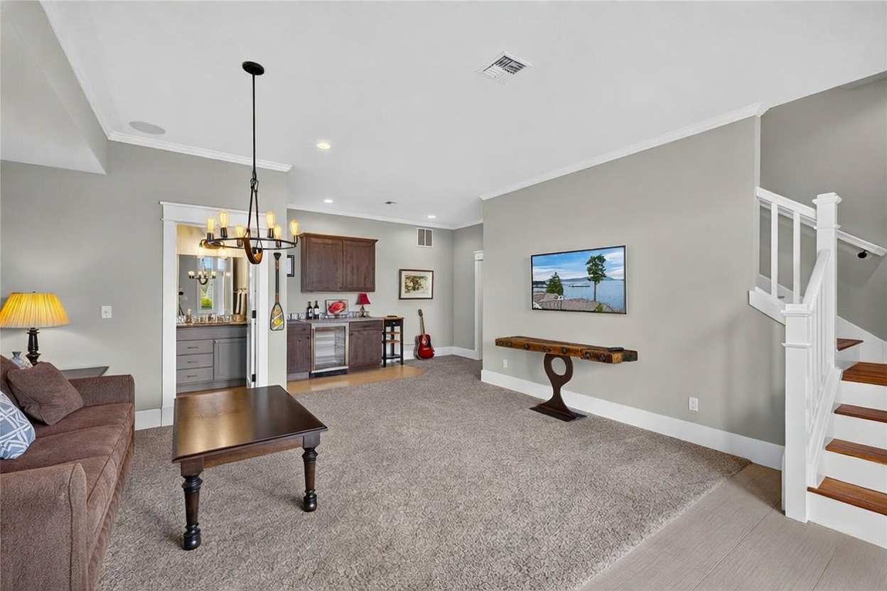 This spacious living area features neutral gray walls, plush carpeting, and a comfortable brown sofa paired with a dark wood coffee table. A wet bar with dark cabinetry and a built-in wine cooler is tucked into an alcove, while a wall-mounted television sits above a unique, rustic-style console table. The room is illuminated by a decorative chandelier and includes a staircase with white railings leading to an upper level.