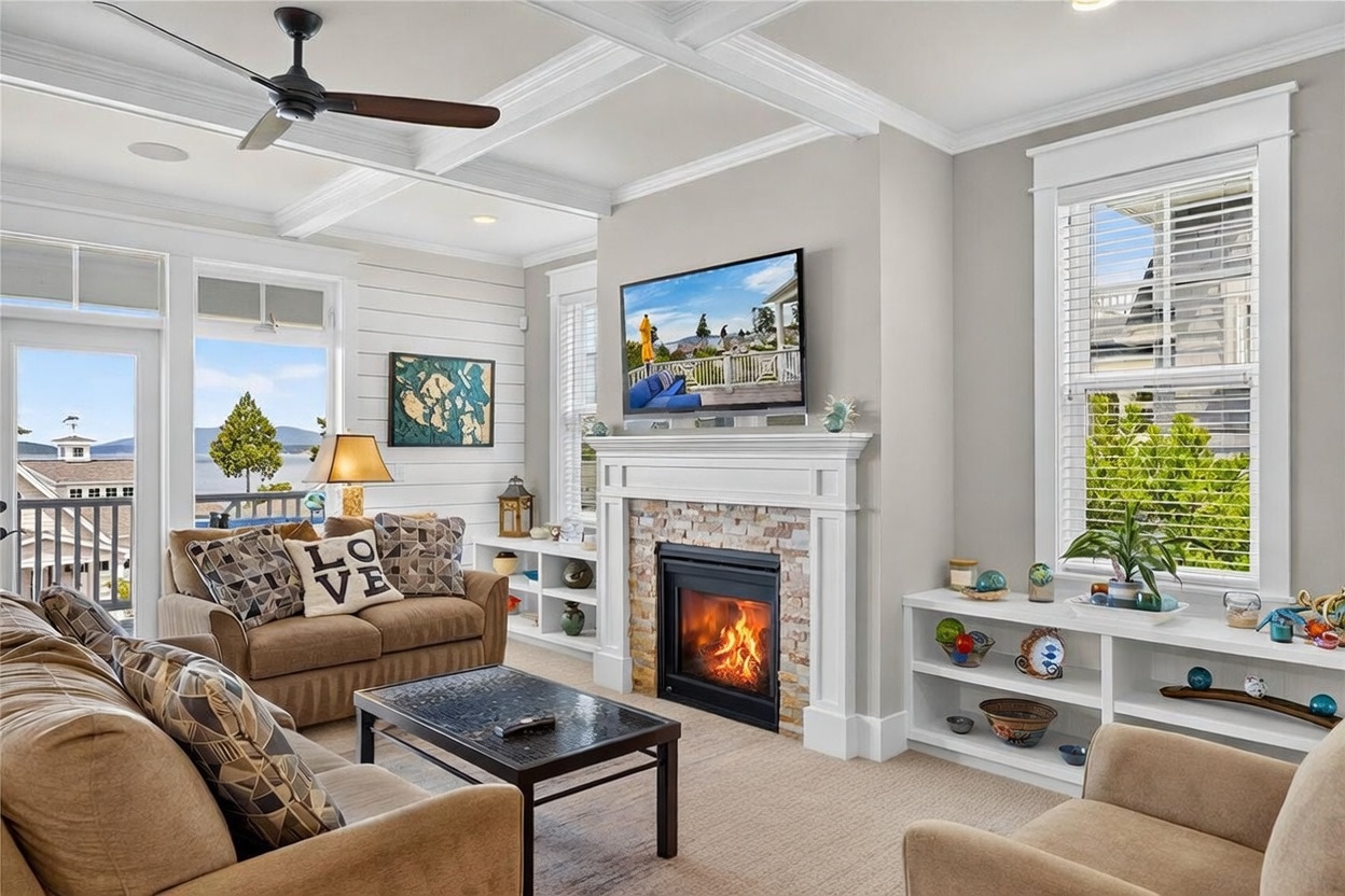 This inviting living room features a cozy seating area centered around a fireplace with a stone-clad chimney breast and a white mantel. The space is brightened by large windows and a glass door that provides a scenic view of the water, while a coffered ceiling adds architectural interest. Neutral-toned furniture, including a plush sofa and a leather ottoman, creates a comfortable and cohesive atmosphere.