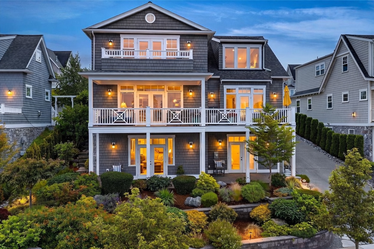 This three-story coastal-style home features dark gray shingle siding and a prominent white-railed balcony spanning the width of the second level. The rear facade includes multiple sets of glass doors and large windows that illuminate the interior, while a lush, tiered garden with stone retaining walls leads up to the ground-level patio. The scene is captured at dusk, with warm exterior lighting creating an inviting and sophisticated atmosphere.