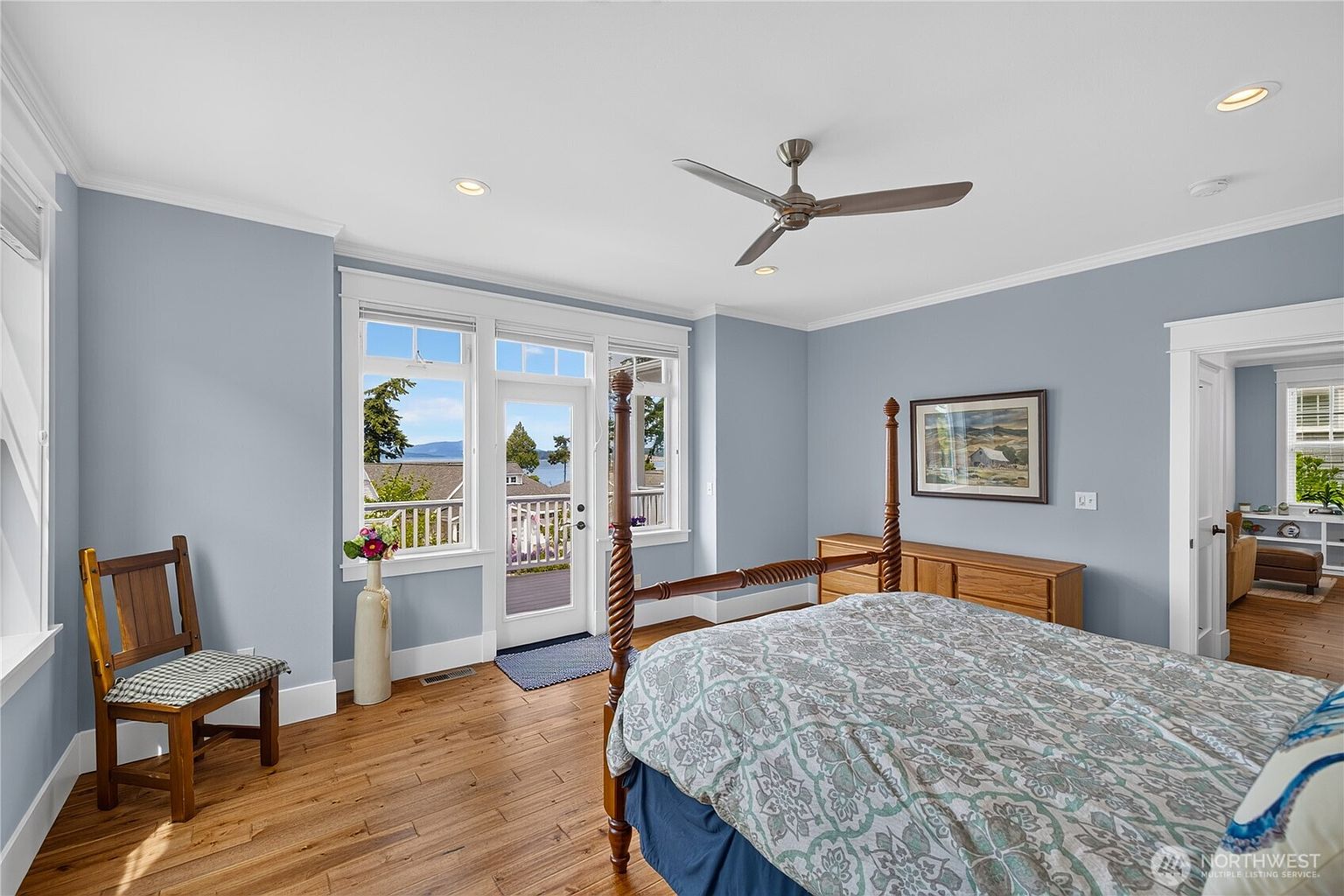This primary bedroom features light blue walls, warm-toned wood flooring, and a four-poster bed as the central focal point. A glass door flanked by large windows provides access to a balcony with scenic water views, filling the room with natural light. The space is accented by a wooden dresser, a decorative chair, and a framed landscape painting, creating a serene and inviting atmosphere.