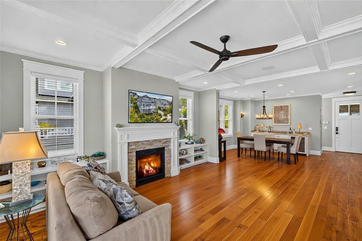 This inviting living room features a coffered ceiling and a central fireplace with a stone-clad surround and a classic white mantel. The space is furnished with two plush, neutral-toned sofas arranged around a dark coffee table, creating a comfortable seating area. Beyond the living room, an open-concept dining area and a wet bar are visible, showcasing a cohesive and well-lit floor plan.