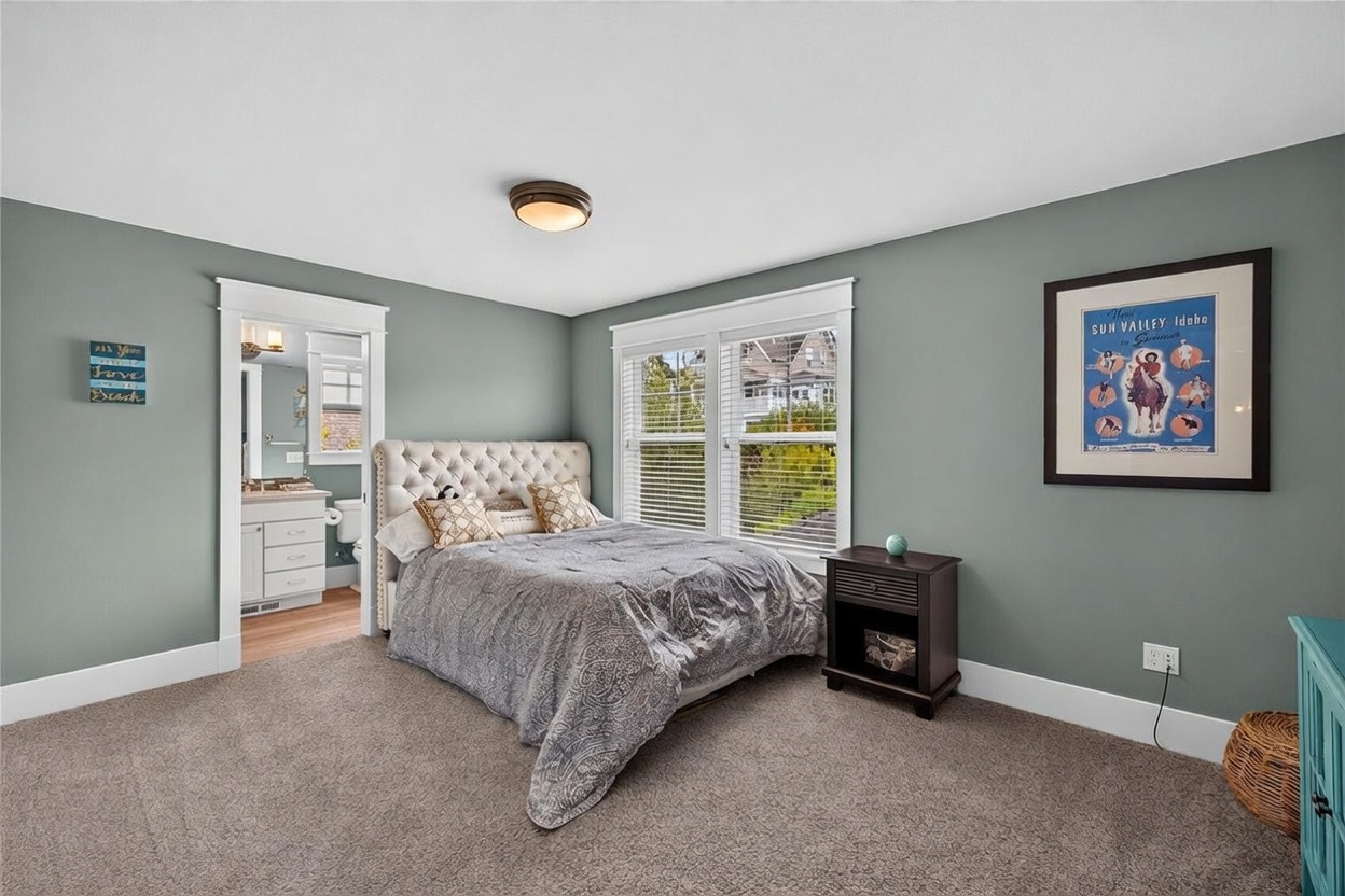 This cozy bedroom features sage green walls, plush carpeting, and a tufted upholstered headboard on the bed. A dark wood nightstand sits beside the bed, while a doorway leads directly into an adjoining bathroom. Large windows with white blinds allow natural light to fill the space, creating a bright and inviting atmosphere.
