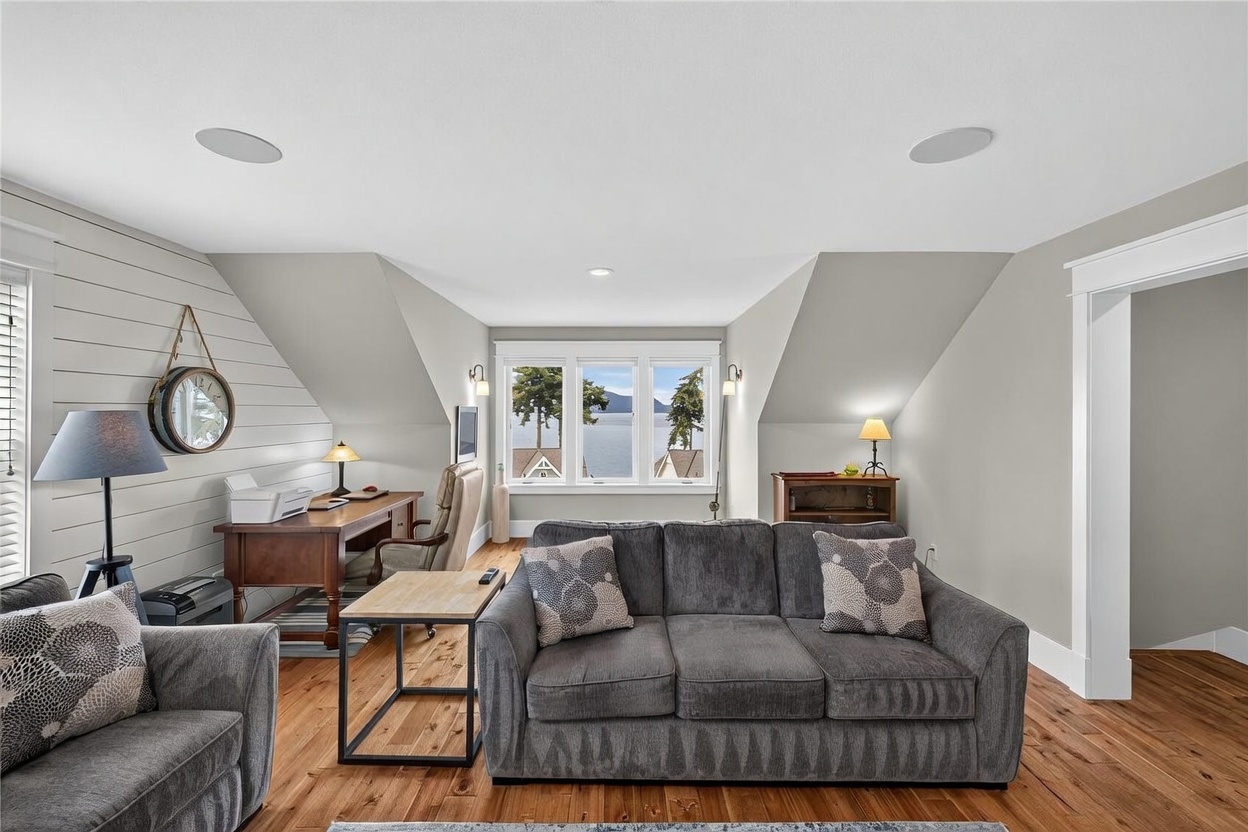 This multi-functional room serves as both a cozy living area and a home office, featuring warm wood flooring and a light-toned shiplap accent wall. A large gray sofa sits in the foreground, while a wooden desk and office chair are positioned near a set of windows that provide a scenic view of the water. The space is bright and inviting, with recessed lighting and a neutral color palette that creates a comfortable atmosphere.