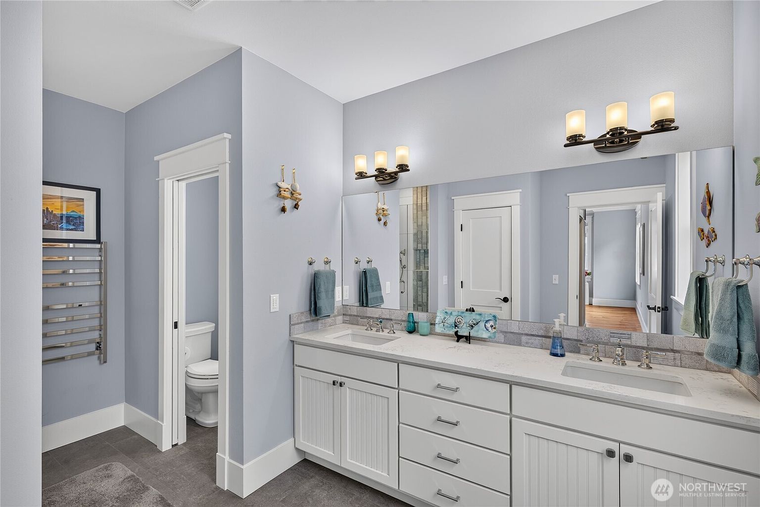 This bright and airy primary bathroom features a double vanity with white cabinetry, a light-toned stone countertop, and dual sinks. A large, wide mirror spans the wall above the vanity, reflecting the light-blue walls and the doorway leading to a private water closet. The space is finished with dark tile flooring and includes a wall-mounted towel warmer, creating a clean and functional aesthetic.