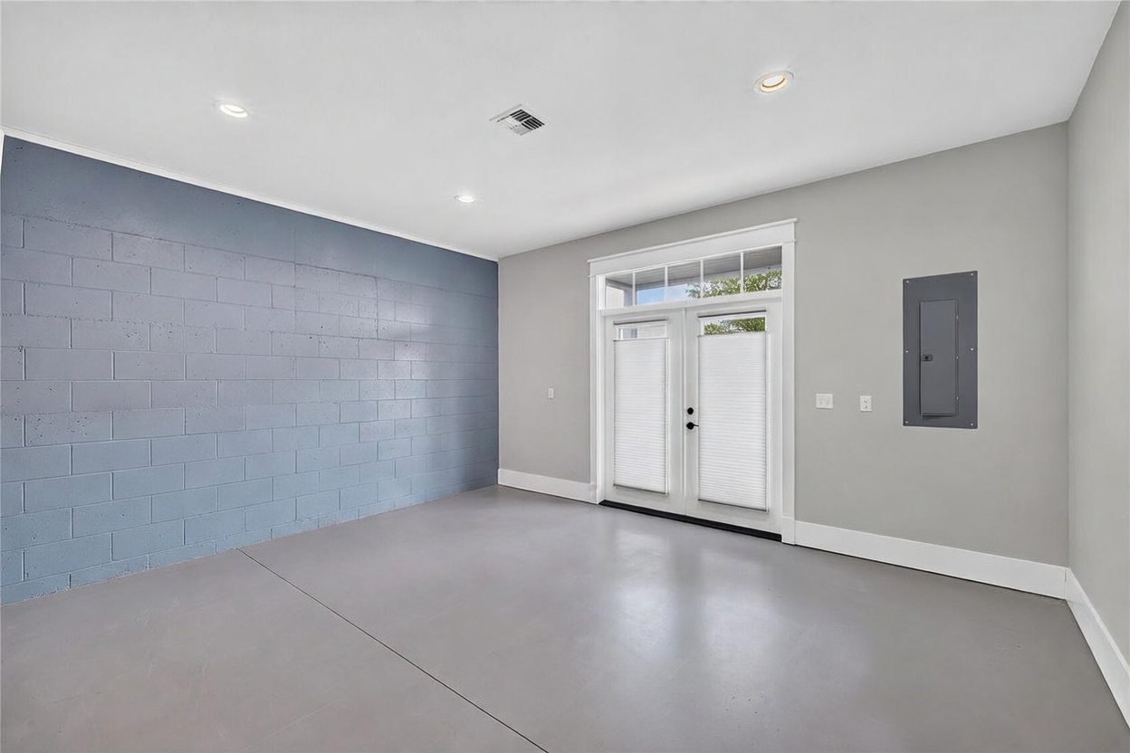 This interior space features a clean, minimalist aesthetic with a smooth, light-colored floor and a distinctive blue-painted concrete block accent wall. A set of double glass doors with white frames and integrated blinds provides natural light, while recessed lighting is installed throughout the ceiling. The room includes a wall-mounted electrical panel, suggesting a functional space such as a finished garage or a versatile flex room.